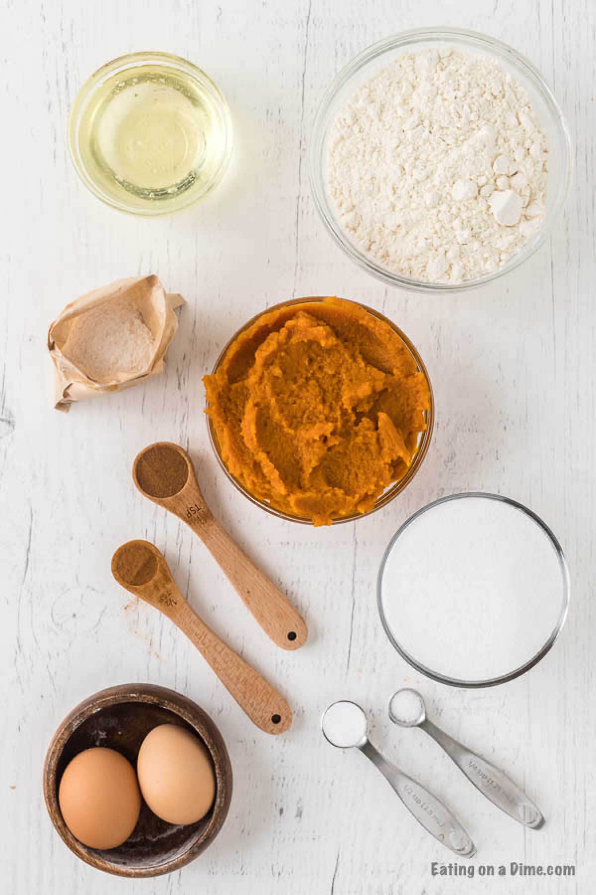 Various baking ingredients are arranged on a white surface: a bowl of flour, sugar, and pumpkin puree—the essentials for a delightful pumpkin bread recipe—alongside two eggs, oil, brown sugar, and measuring spoons with baking powder and spices.