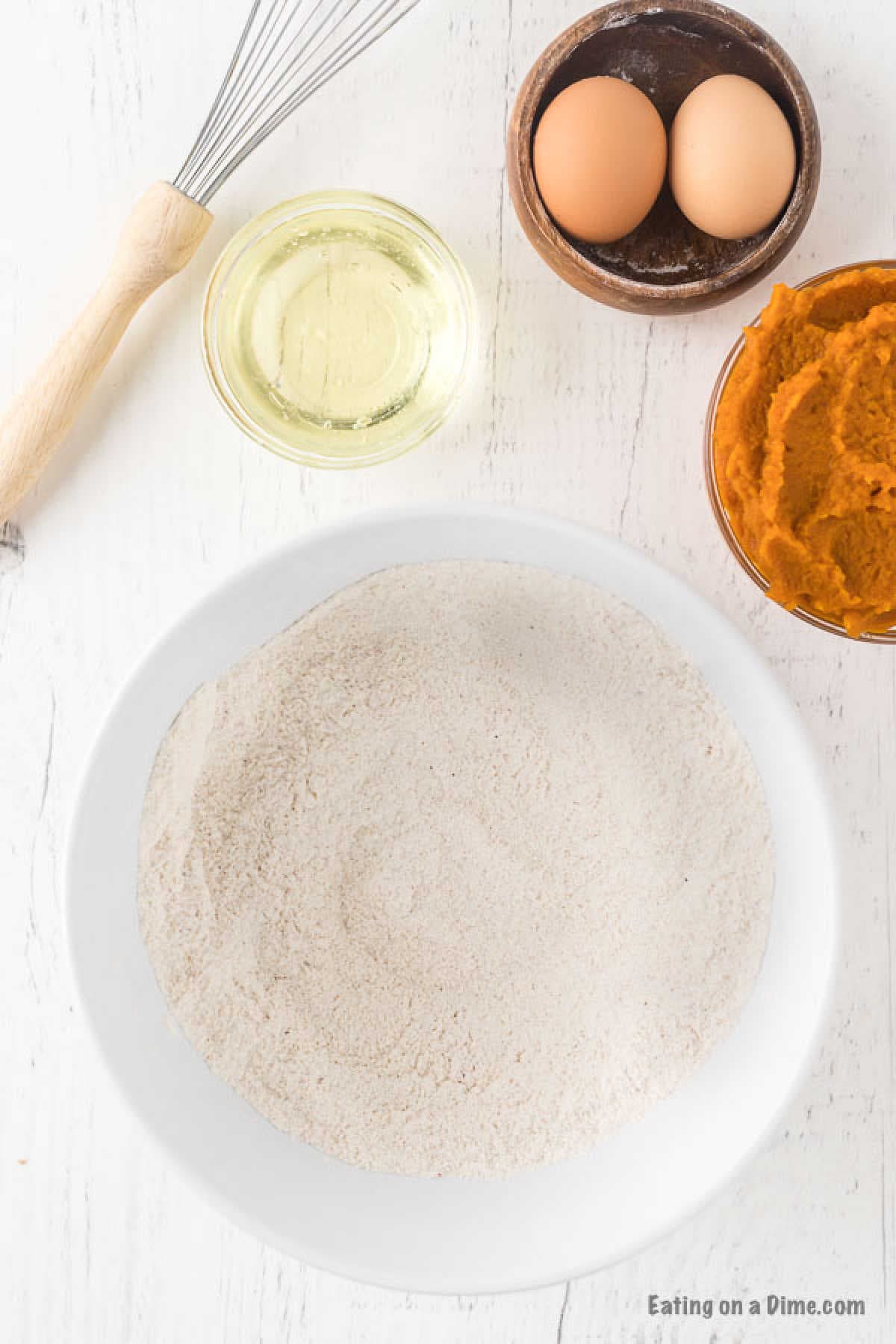 A white bowl with flour sits on a white surface, ready for the perfect pumpkin bread recipe. Surrounding it are a whisk, two eggs in a small bowl, a jar of clear liquid, and a bowl of orange puree.