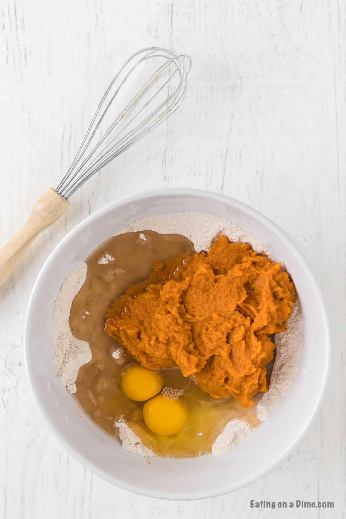 A white bowl contains a mixture of flour, two cracked eggs, pumpkin puree, and a liquid ingredient—a perfect start to a pumpkin bread recipe. A whisk with a wooden handle rests beside the bowl on a white surface.