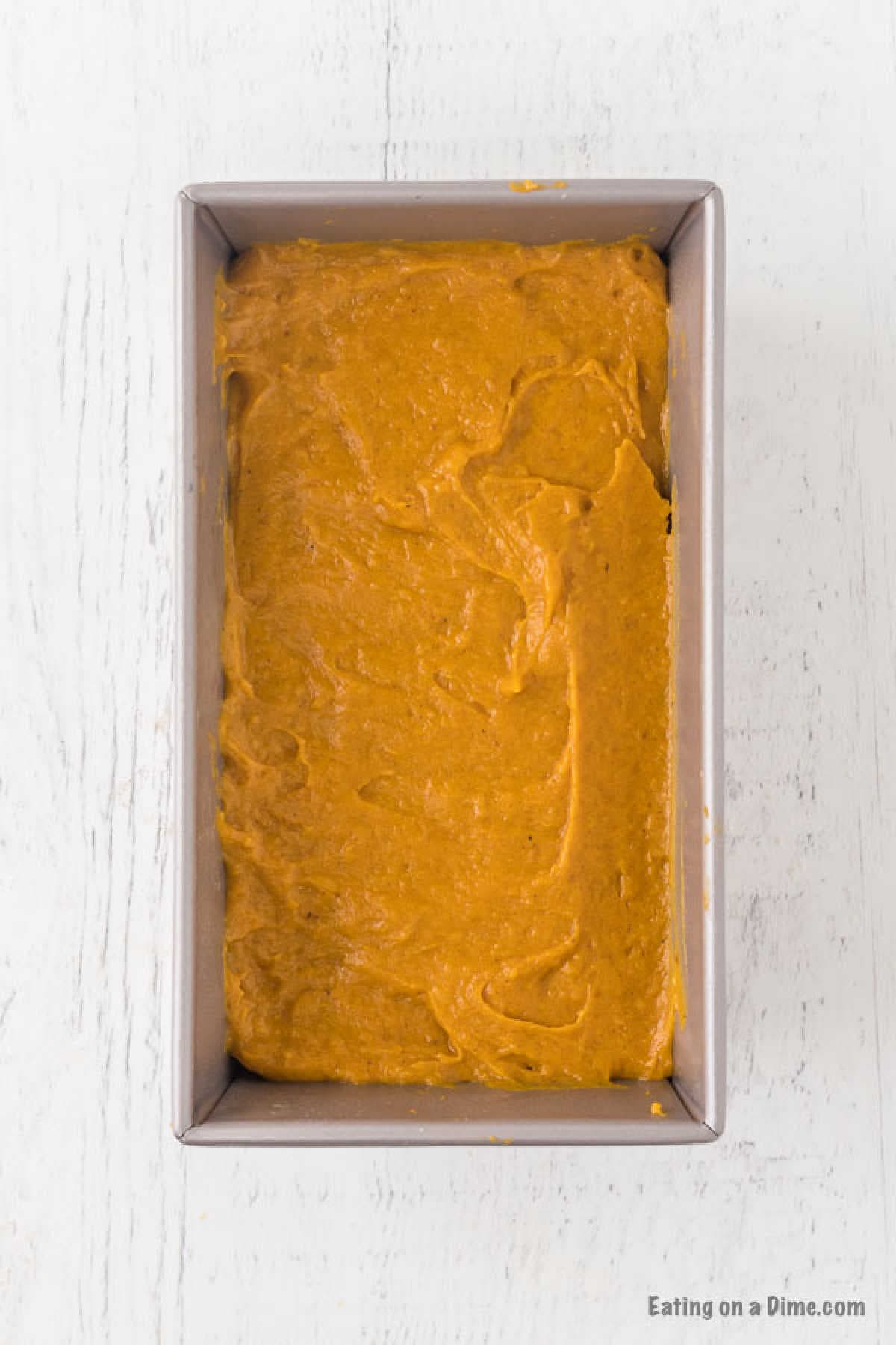 A rectangular baking pan filled with smooth, unbaked pumpkin bread batter from a delightful pumpkin bread recipe sits on a white wooden surface. The batter is evenly spread, showcasing a rich, orange-brown color that promises delicious warmth and flavor.