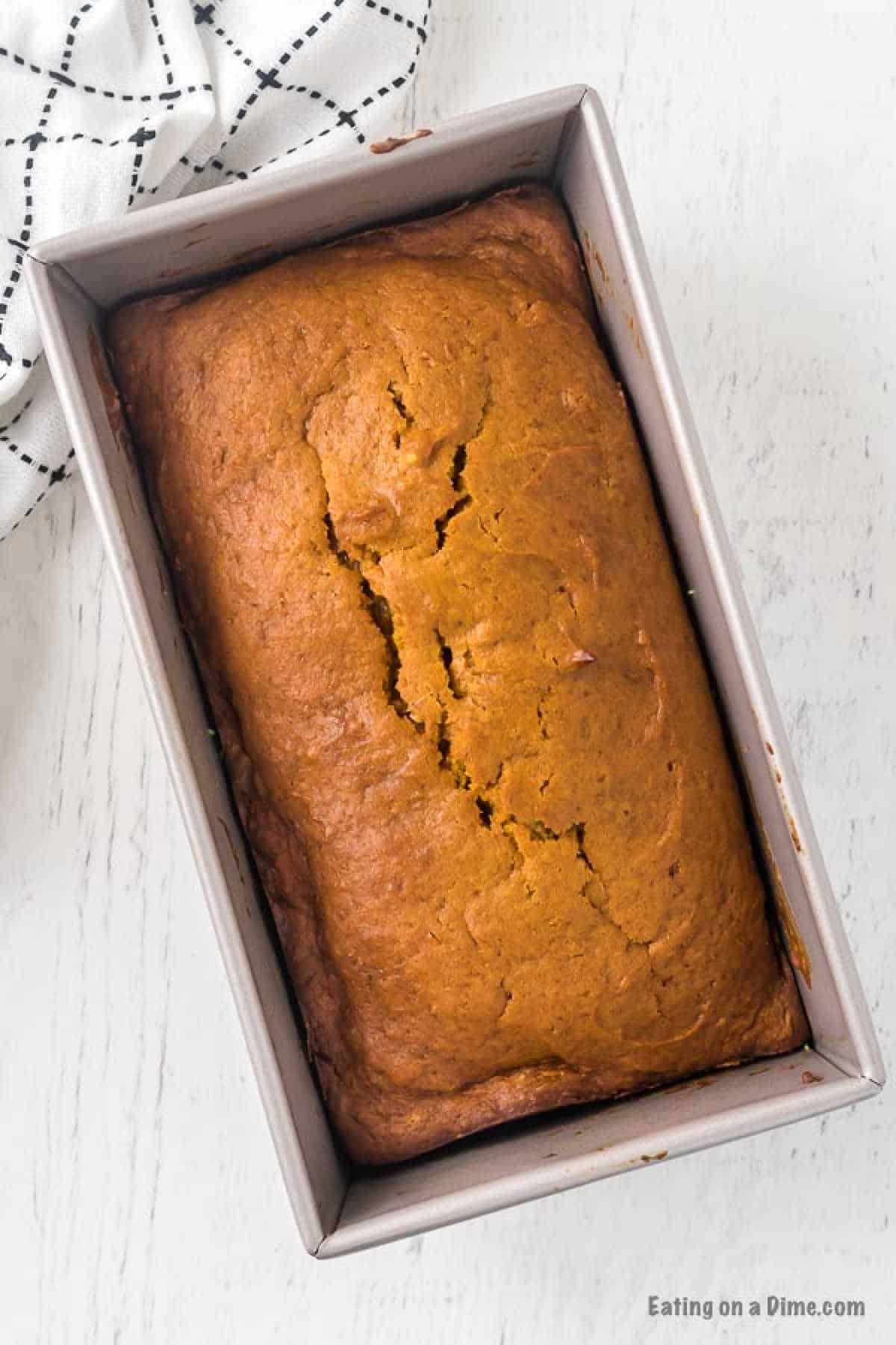 A loaf of freshly baked pumpkin bread rests in a rectangular pan on a white surface, as if straight from a cherished pumpkin bread recipe. A checkered black and white cloth accents the scene's rustic charm, while the golden-brown crust boasts a delightful crack on top.