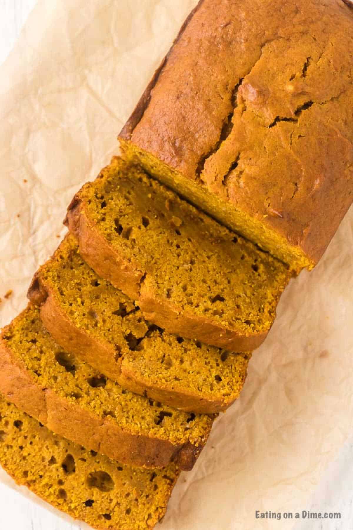 A loaf of pumpkin bread with three slices cut and laid out on a crumpled piece of parchment paper. The bread, inspired by a classic pumpkin bread recipe, boasts a golden brown crust and moist, rich orange interior, suggesting a spiced, aromatic flavor.