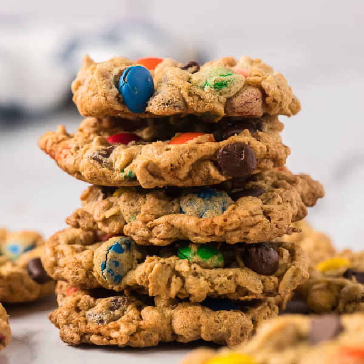 A stack of colorful monster cookies, showcasing an irresistible monster cookie recipe with M&M's and chocolate chips, sits on a marble surface. Blurred cookies are visible in the background.