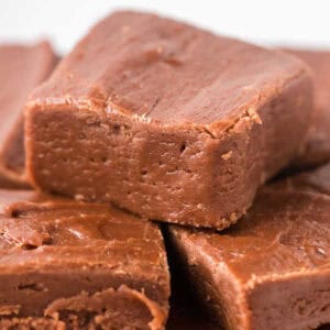 Close-up of several pieces of chocolate fudge stacked on top of each other. The fudge, like an ideal example for those learning how to make fudge, boasts a smooth, glossy surface and a rich, dark brown color. Small cracks and texture detail enhance the creamy appearance.