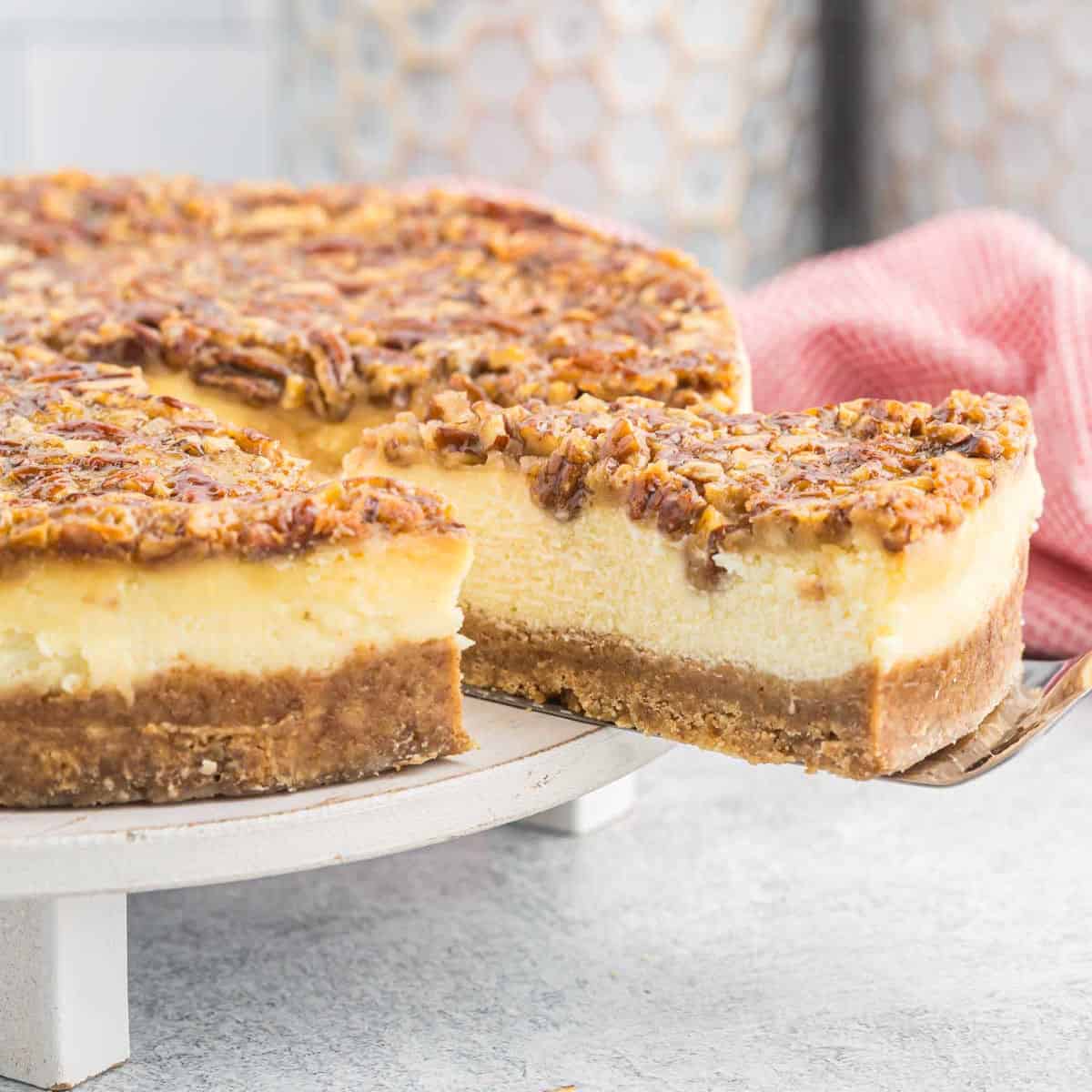 A rich pecan pie cheesecake sits on a white cake stand. One slice is partially pulled out, revealing layers of crumbly crust, creamy cheesecake, and pecan topping. A red and white checkered cloth is blurred in the background.