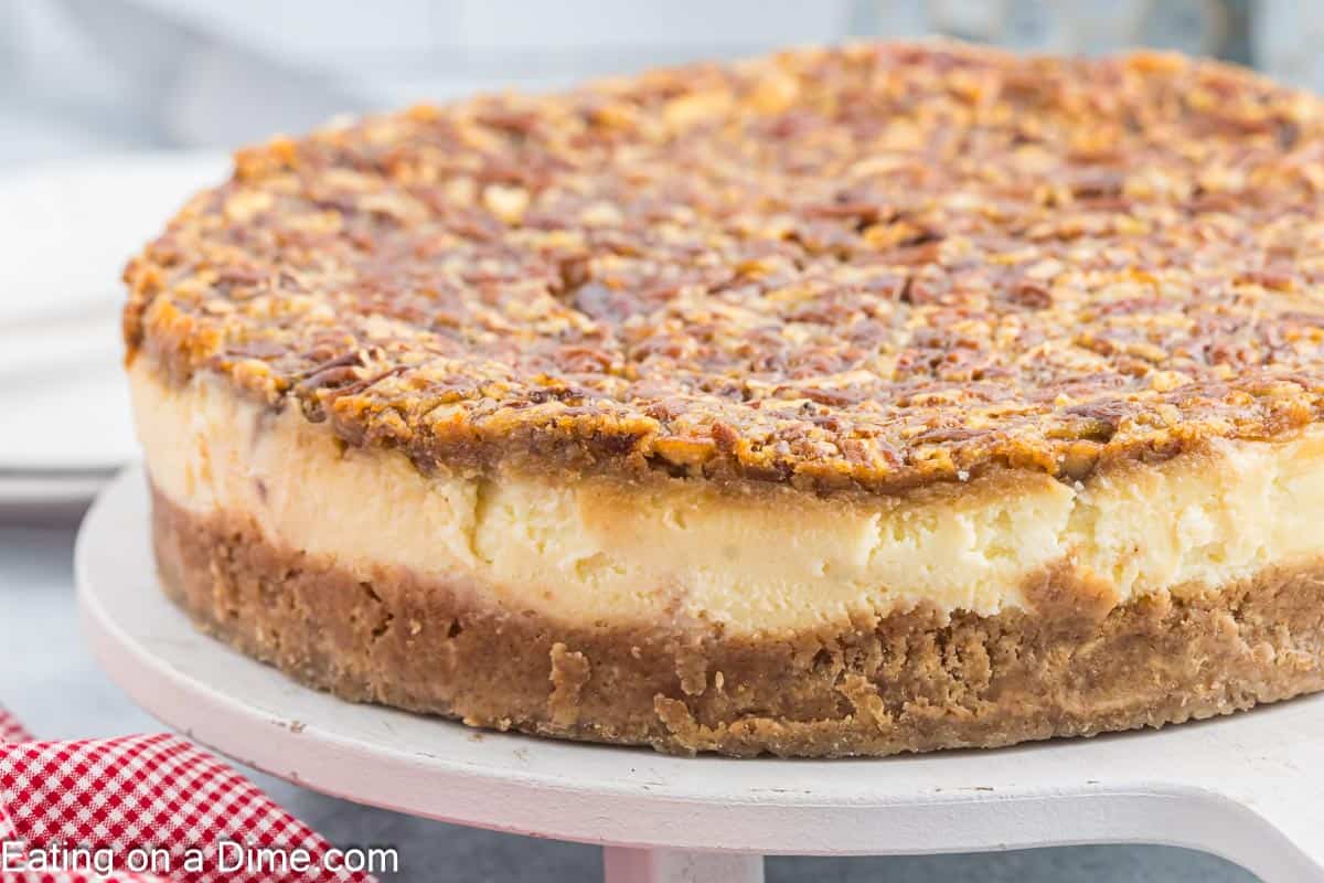 A whole Pecan Pie Cheesecake sits on a white cake stand, featuring a dense, crumbly base with a golden brown, nutty topping. The smooth, creamy middle layer is sandwiched between the crust and the top layer. A red and white checkered cloth is partially visible in the bottom left corner.