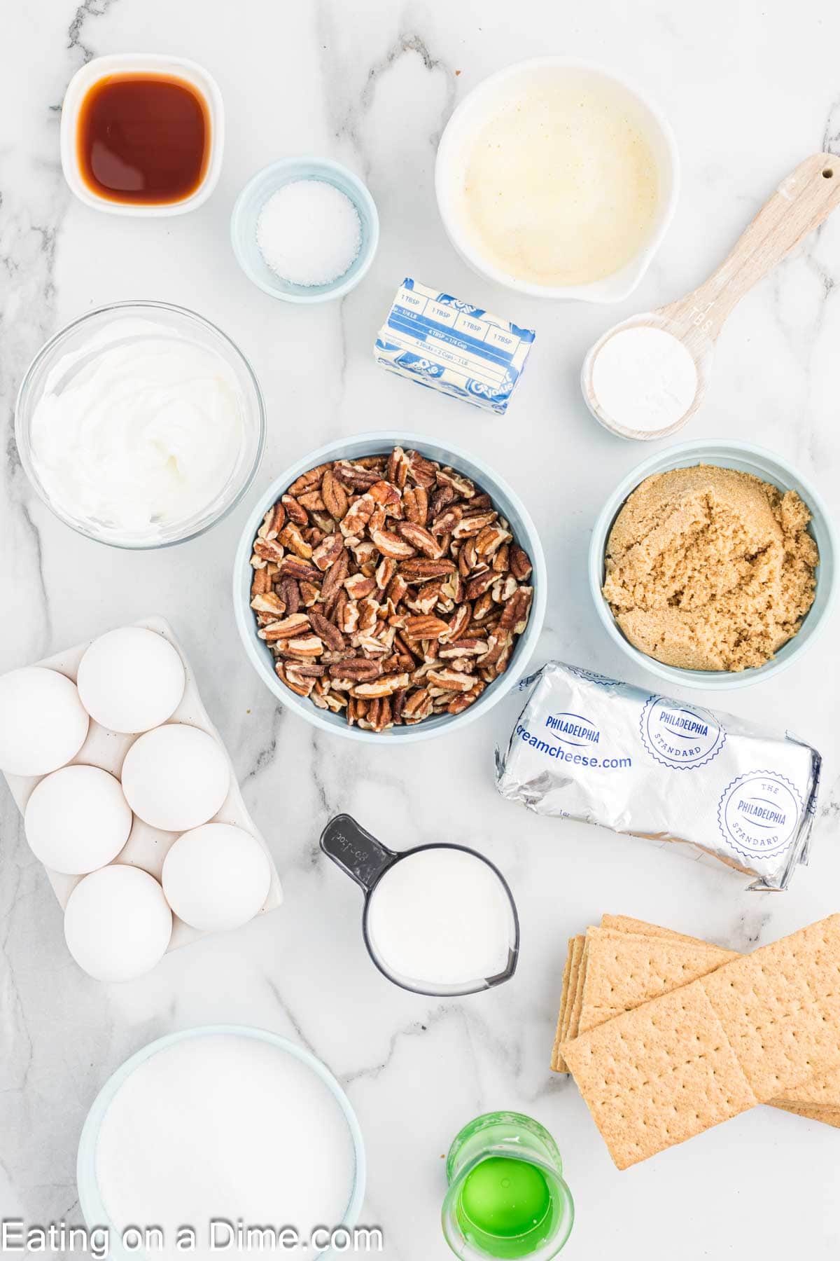 A marble countertop with various baking ingredients laid out, perfect for making a Pecan Pie Cheesecake. It includes a bowl of pecans, brown sugar, cream cheese packets, graham crackers, a small pitcher of milk, eggs, sugar, yogurt bowl, vanilla extract, butter sticks, salt shakers, cornstarch packets, sour cream tubs and a green bottle.