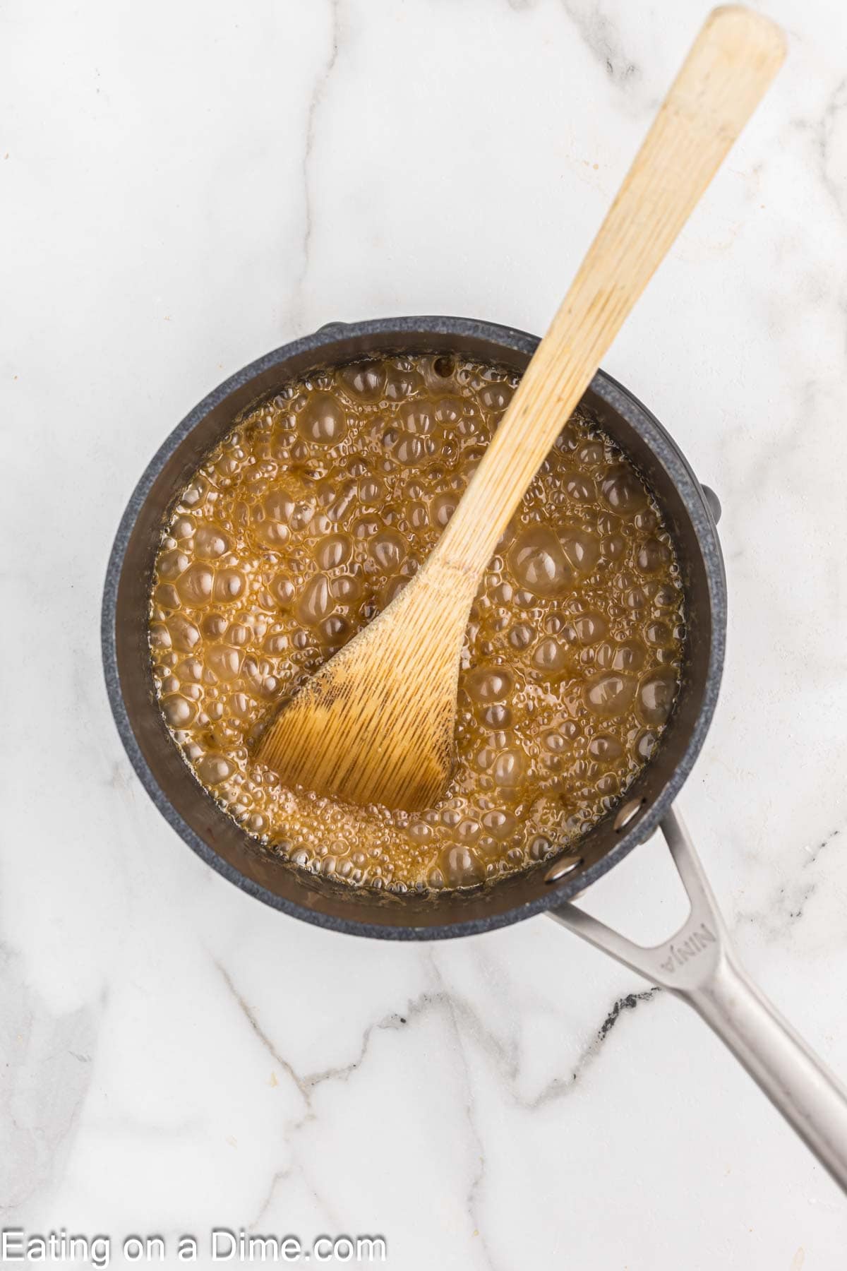 A small black saucepan filled with bubbling caramel sauce sits on a white marble surface, reminiscent of pecan pie cheesecake. A wooden spoon is placed in the sauce, appearing to stir it. The handle of the saucepan extends to the right side of the image.