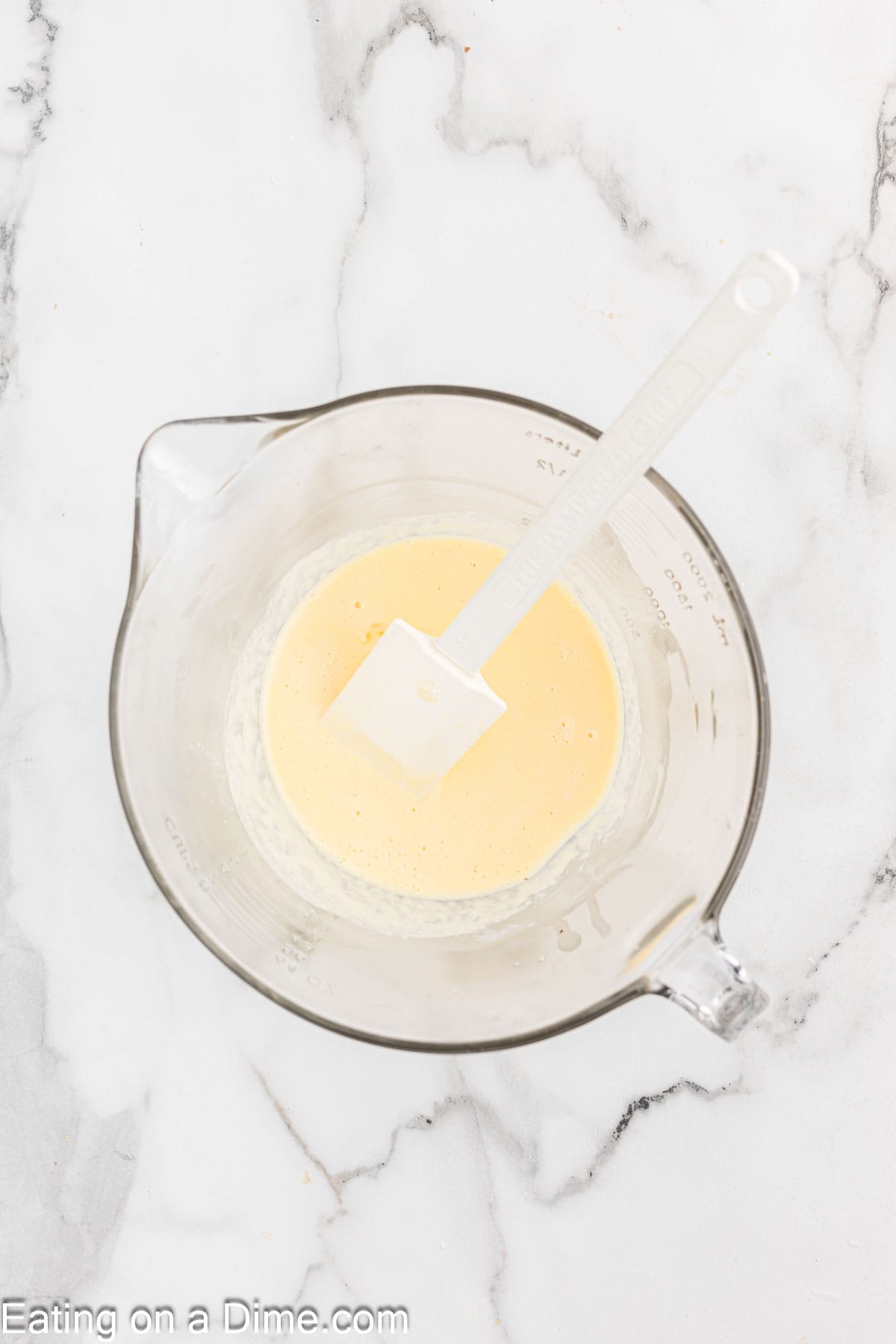 A glass measuring cup filled with a creamy yellow mixture and a white spatula inside, set on a marbled countertop. The image has the text "Eating on a Dime.com" at the bottom, suggesting it's an ingredient for their delectable Pecan Pie Cheesecake.