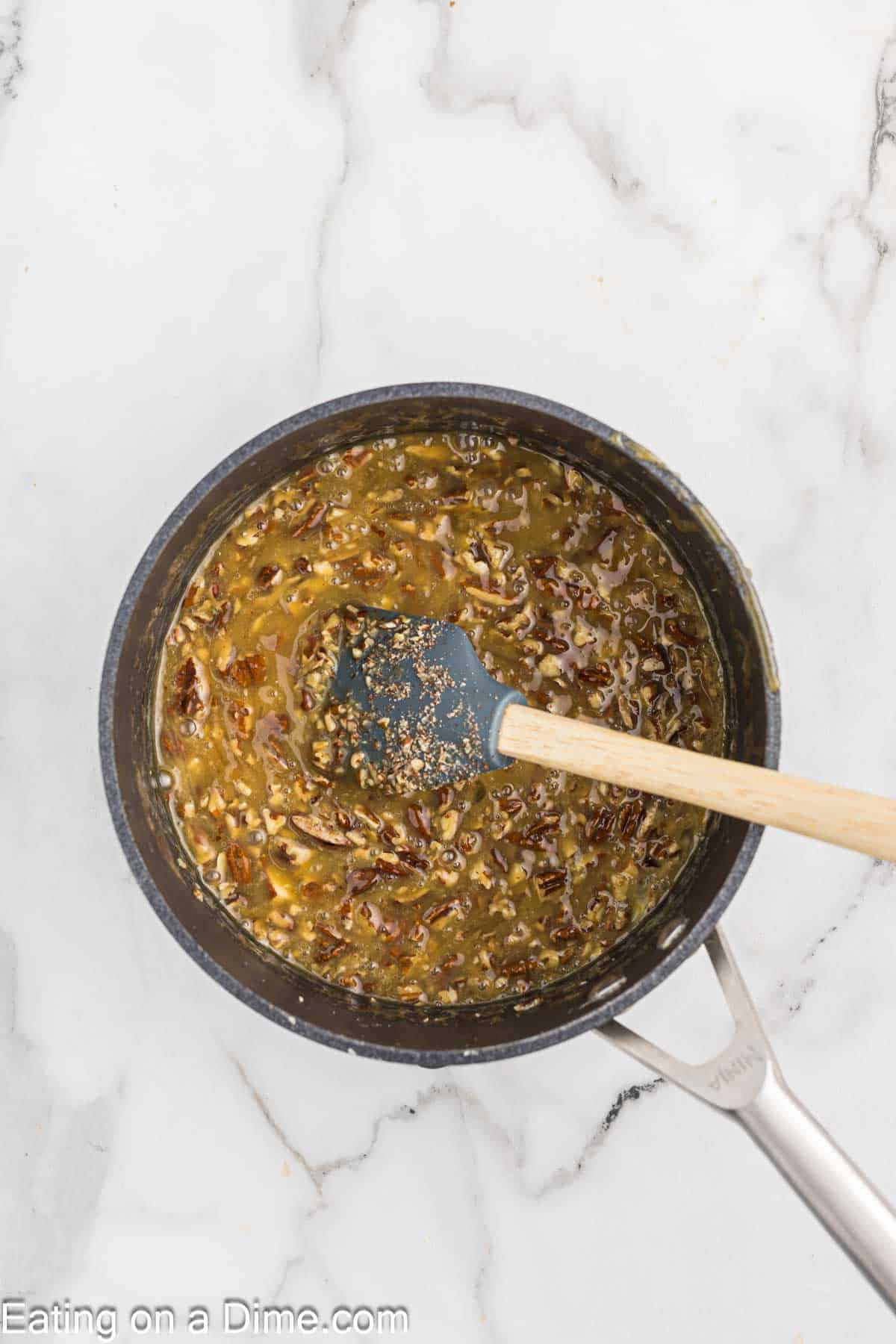 A saucepan filled with a mixture of chopped nuts and a caramel-like sauce, reminiscent of pecan pie cheesecake, is being gently stirred with a wooden-handled silicone spatula. The pan rests elegantly on a white marble surface.