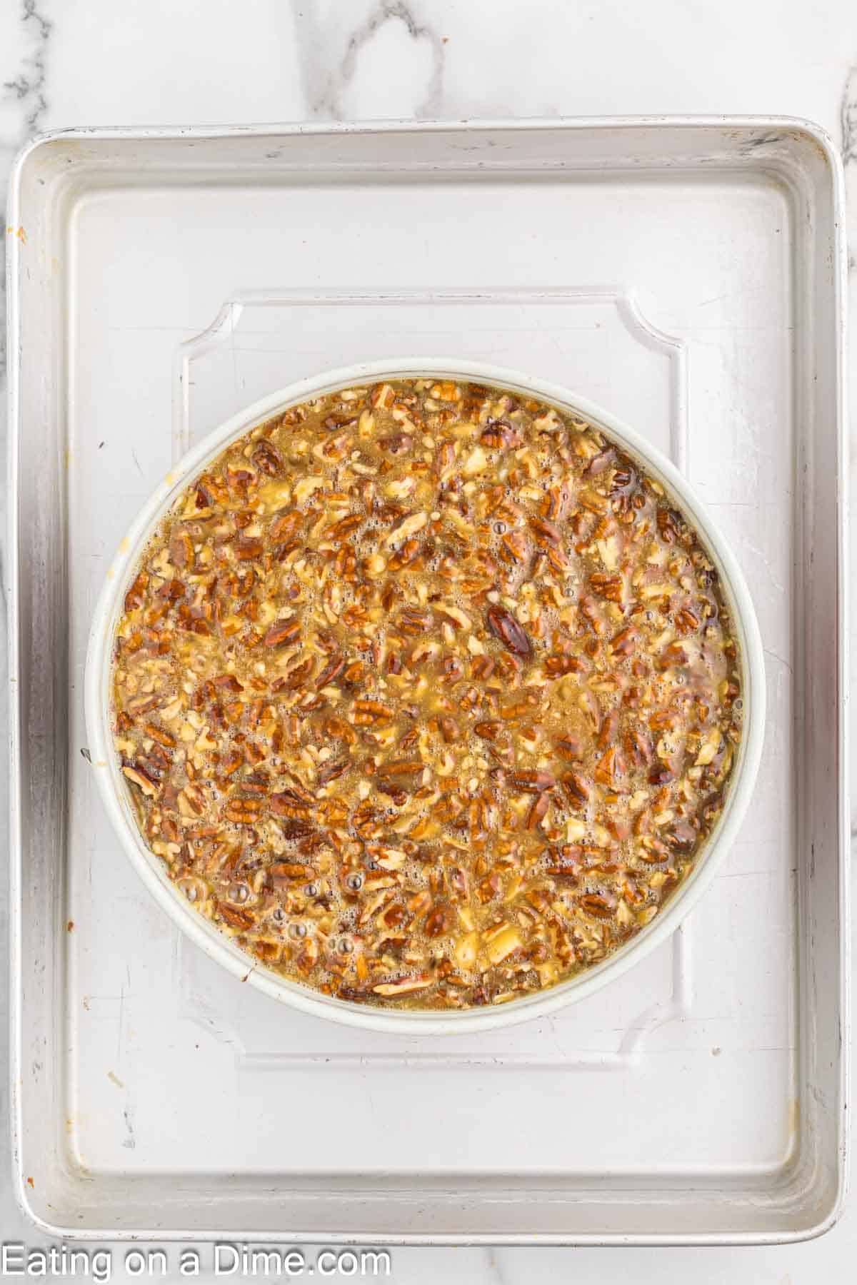 A round baking dish filled with a mixture resembling pecan pie filling, placed on a silver baking tray. The decadent mix, akin to Pecan Pie Cheesecake, consists of visible chopped pecans in a caramel-colored blend. The white text "Eating on a Dime.com" is at the bottom of the image.