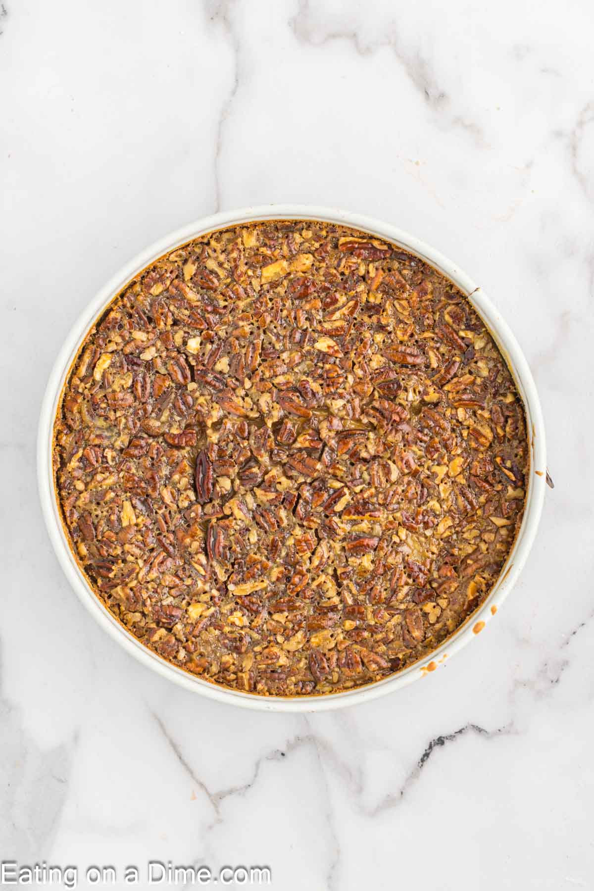 A round Pecan Pie Cheesecake sits on a white marble surface, showcasing a rich, nutty top layer with a golden-brown crust.