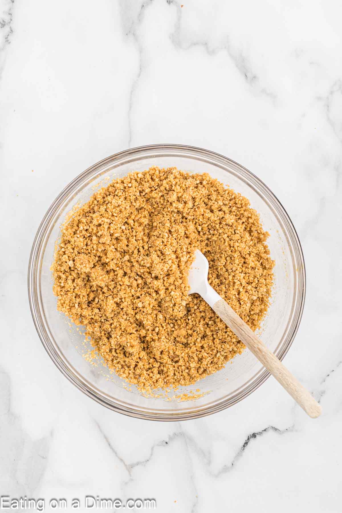 A clear glass bowl filled with a crumbly, golden brown mixture sits on a white marble countertop. A wooden spoon with a white handle rests inside the bowl, evoking thoughts of Pecan Pie Cheesecake. The website "Eating on a Dime.com" is visible at the bottom of the image.