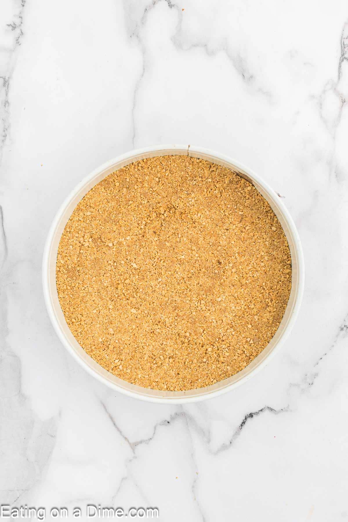 A bowl filled with ground graham cracker crumbs on a white marble surface. The background shows a subtle pattern in the marble. At the bottom of the image, partially visible text reads, "Eating on a Dime.com." Perfect for making Pecan Pie Cheesecake!