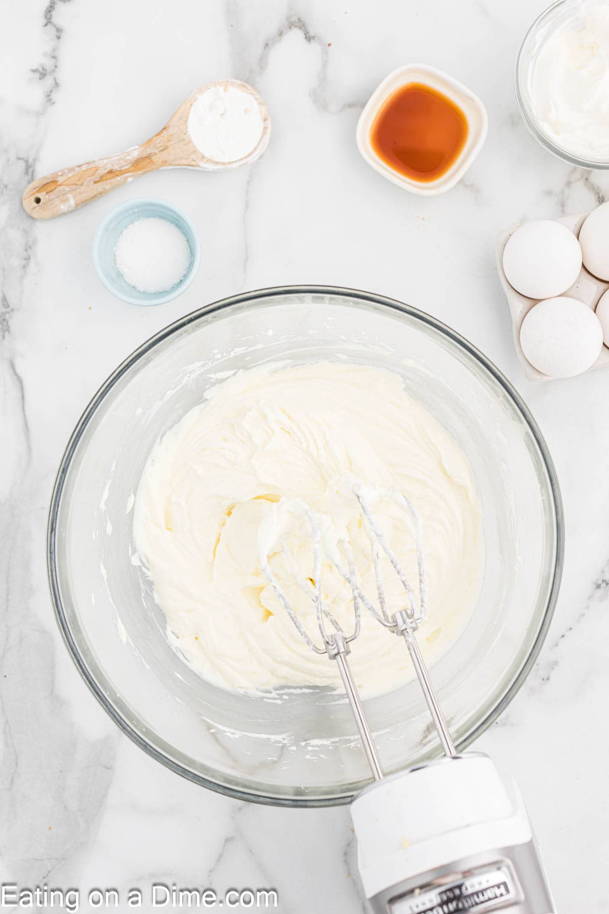 A mixing bowl with creamy batter being mixed by an electric hand mixer is on a marble countertop. Surrounding the bowl are ingredients for Pecan Pie Cheesecake, including eggs, a small bowl of salt, a teaspoon with flour, and a bowl of vanilla extract. “Eating on a Dime.com” is texted at the bottom left.