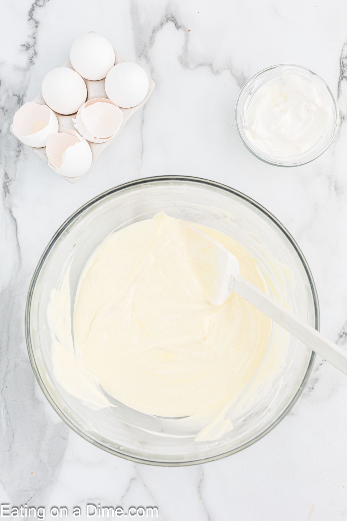 A glass mixing bowl filled with creamy batter is placed on a marble countertop. Beside it are a small glass bowl containing white mixture and an eggshell-filled carton, hinting at the beginnings of a decadent Pecan Pie Cheesecake. The image showcases ingredients and partial preparation for a recipe. "Eating on a Dime.com" watermark is at the bottom.