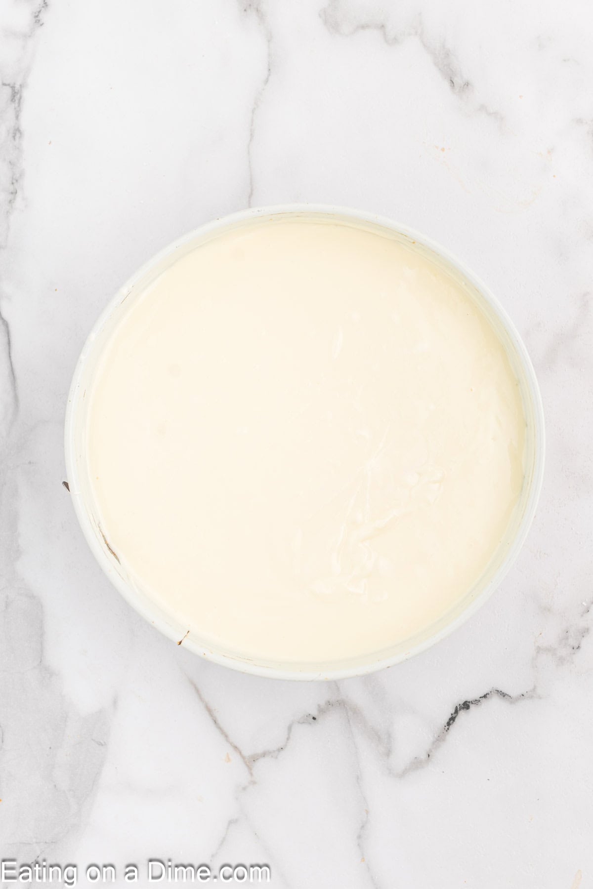 A top-down view of a white bowl containing a smooth, creamy white substance resembling yogurt or a similar dairy product, placed on a white marble countertop. The surface of the creamy substance is even and glossy, evoking the rich texture of Pecan Pie Cheesecake filling.