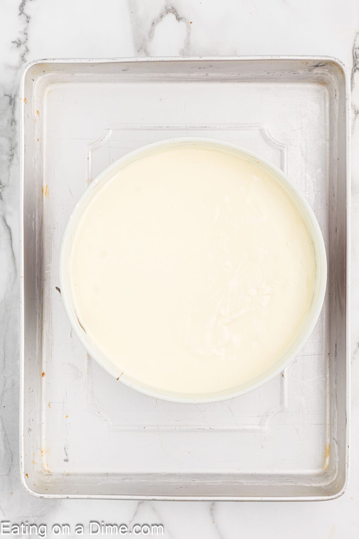 A round white pecan pie cheesecake batter in a circular baking pan placed on a metal baking tray. The tray is set on a white marble surface. The edges of the tray show signs of use, with slight discoloration and marks.