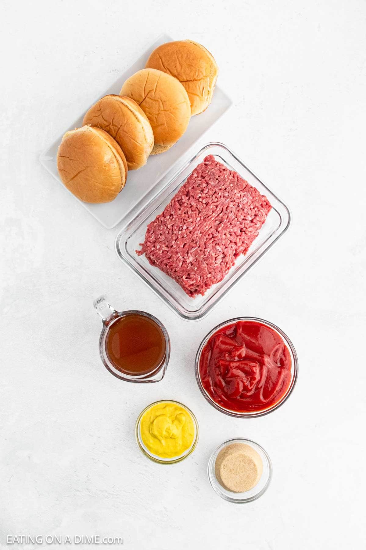 Top-down view of ingredients for Instant Pot sloppy joes on a white background: hamburger buns, a block of ground beef, a cup of broth, a bowl of ketchup, and small bowls of mustard and spices.