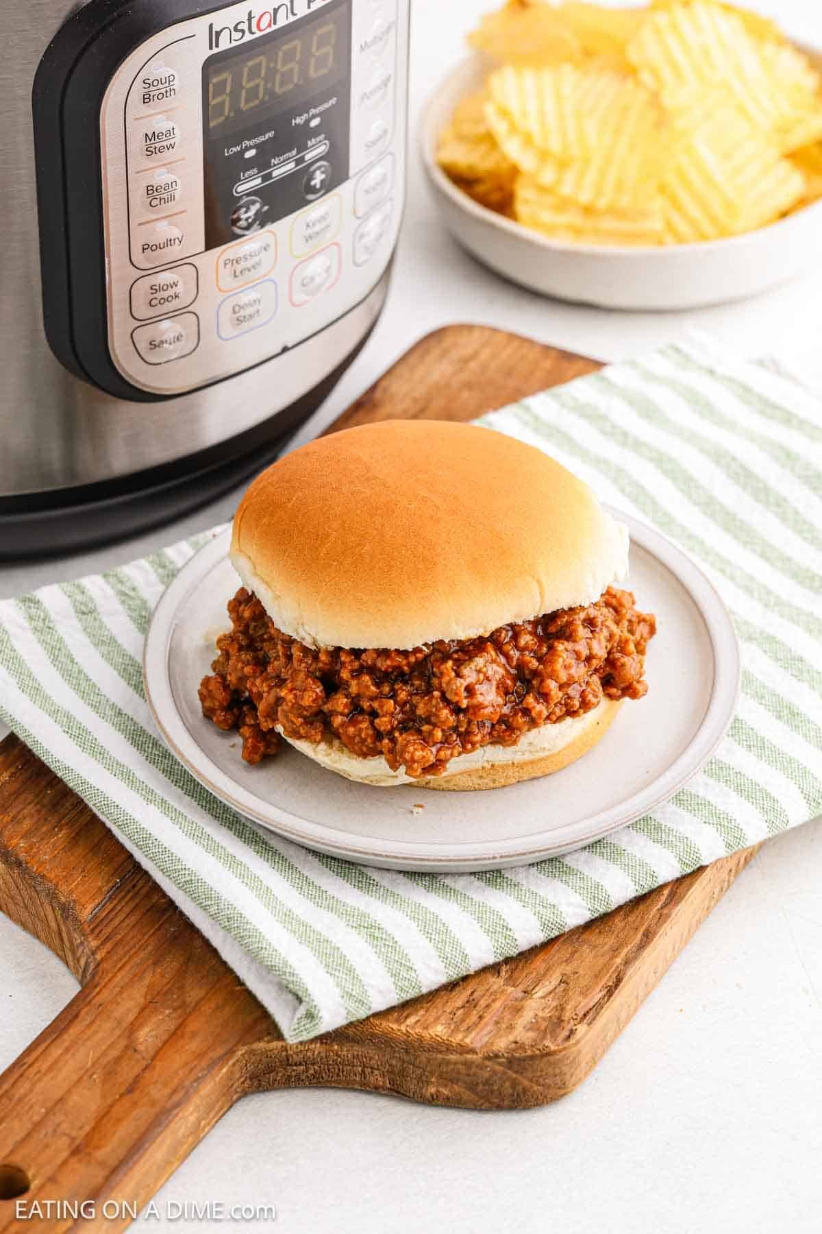 A classic sloppy joe sandwich rests on a striped cloth, with an Instant Pot hinting at the quick preparation in the background. Beside it, potato chips are nestled in a bowl, all set upon a rustic wooden cutting board for a cozy dining experience.