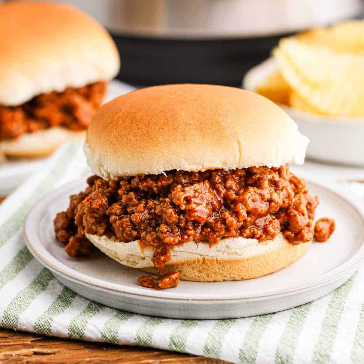 An instant pot sloppy joe sandwich with seasoned ground meat filling on a round bun sits invitingly on a white plate. In the background, there's another sandwich and a bowl of potato chips, all resting on a green and white striped cloth.