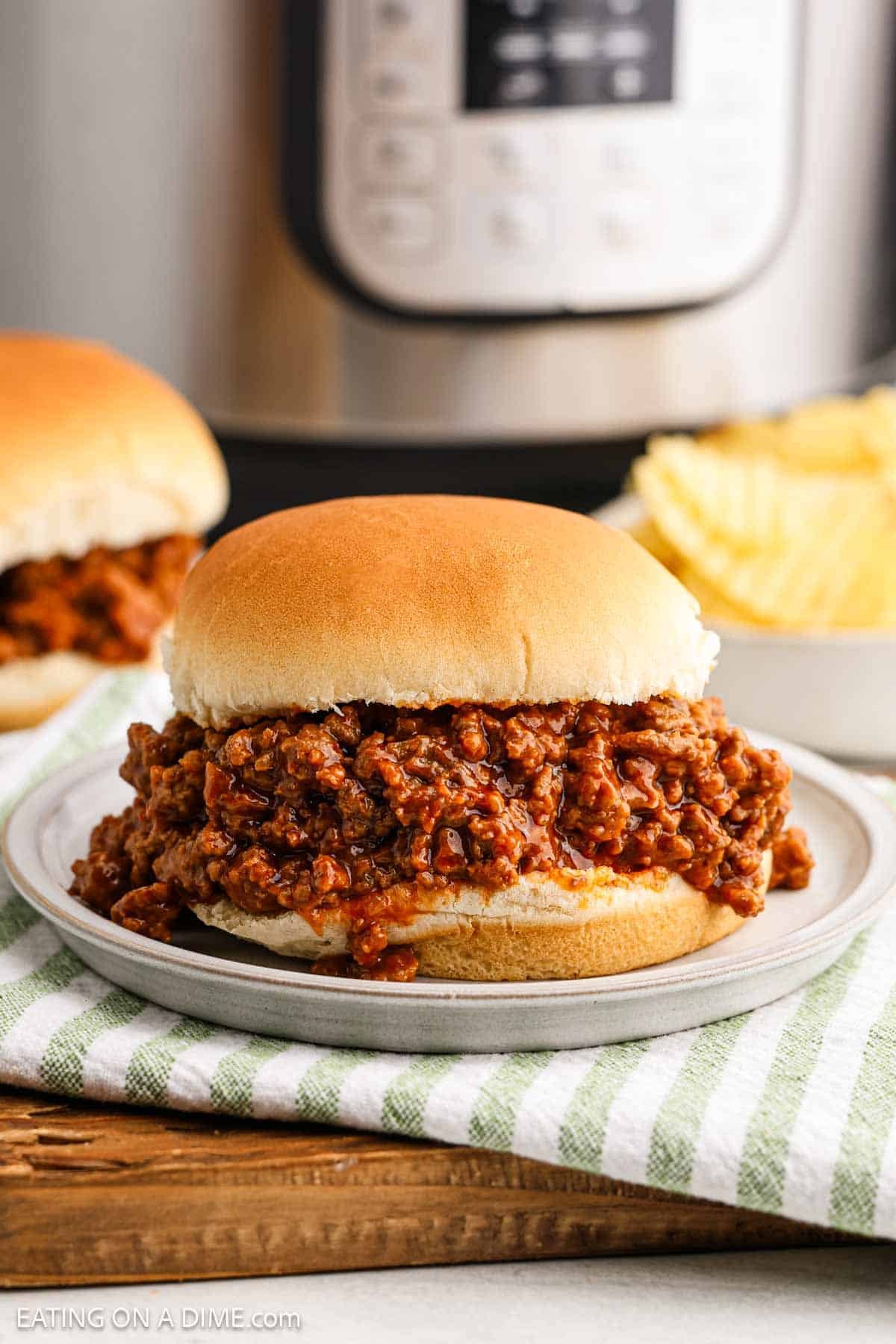 A sloppy joe sandwich brimming with seasoned meat rests on a gray plate, nestled atop a striped cloth. In the background, an instant pot and another sandwich make their appearance, accompanied by a tempting bowl of potato chips.