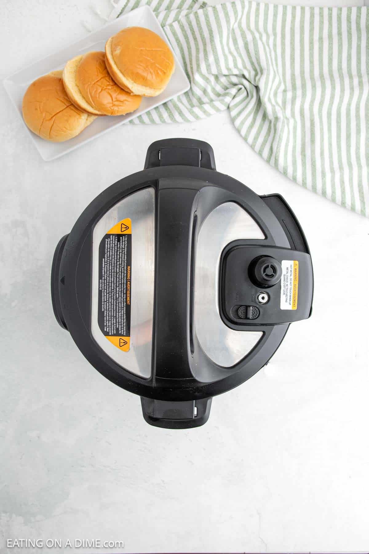 Top view of an Instant Pot with a black lid on a light countertop. A rectangular plate with three burger buns is in the upper left corner, perfect for making sloppy joes, and a green striped cloth is on the right side.