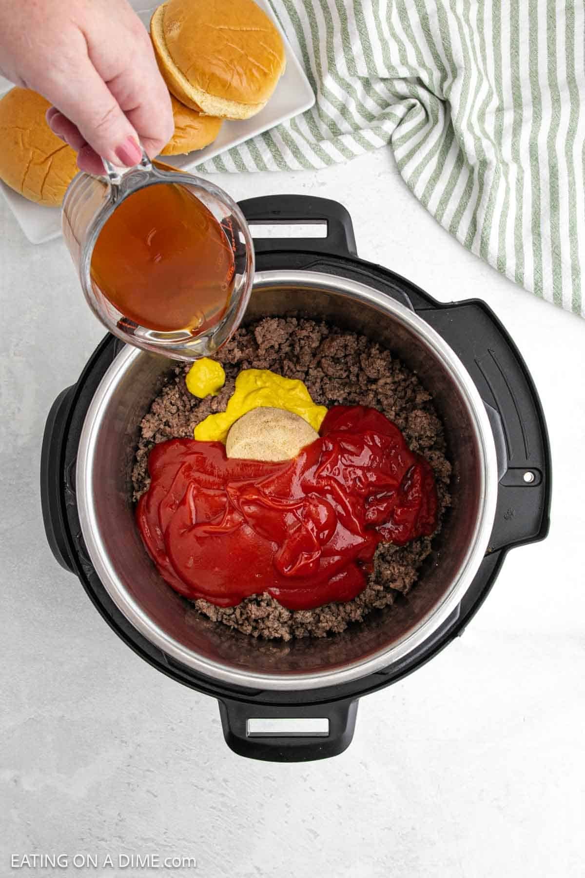 A person pours a liquid into an Instant Pot filled with cooked ground beef, ketchup, mustard, and a brown spice mix—perfect for making delicious Sloppy Joes. Nearby, two burger buns rest on a striped cloth atop a white surface.