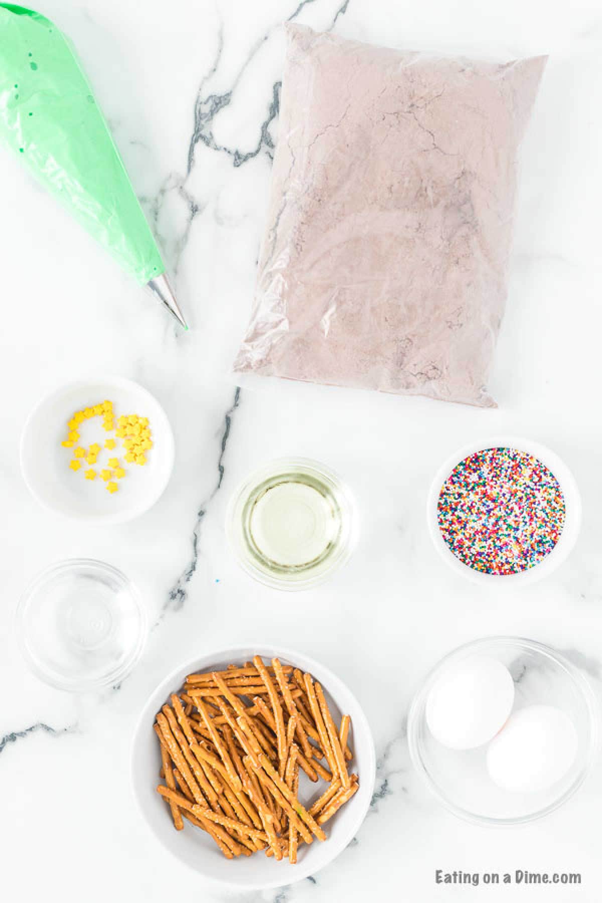 A flat lay image shows various baking ingredients on a white marble surface, perfect for making Christmas Tree Brownies. Items include chocolate powder in a bag, pretzel sticks in a bowl, two eggs, a bowl of colorful sprinkles, a small bowl of yellow candy decorations, and a piping bag with green frosting.