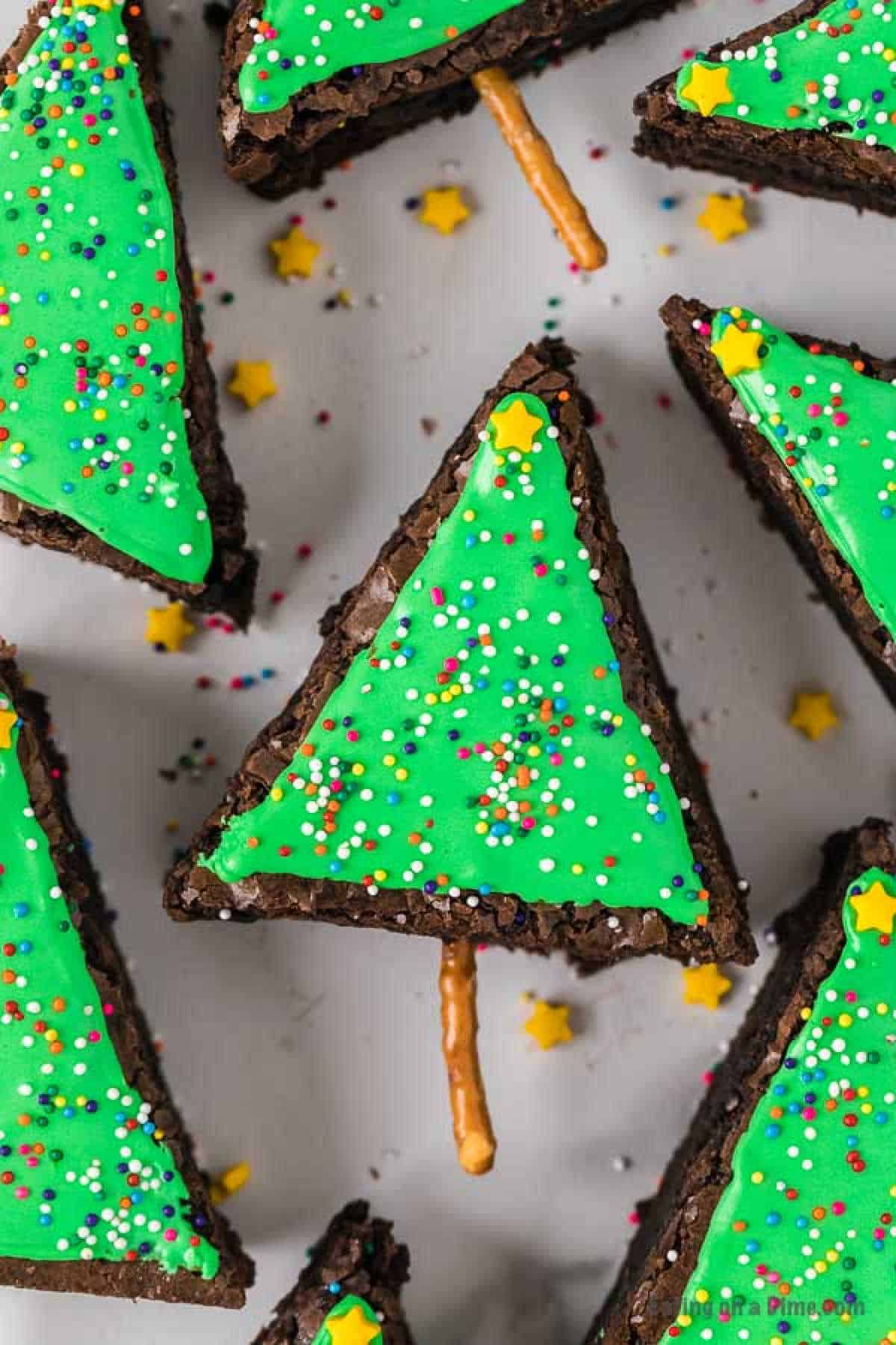 Christmas Tree Brownies are triangular treats decorated with bright green icing and multicolored sprinkles. Each brownie is topped with a small yellow star candy, while a pretzel stick forms the tree trunk. Scattered yellow star candies enhance the festive presentation, making these brownies a holiday delight.