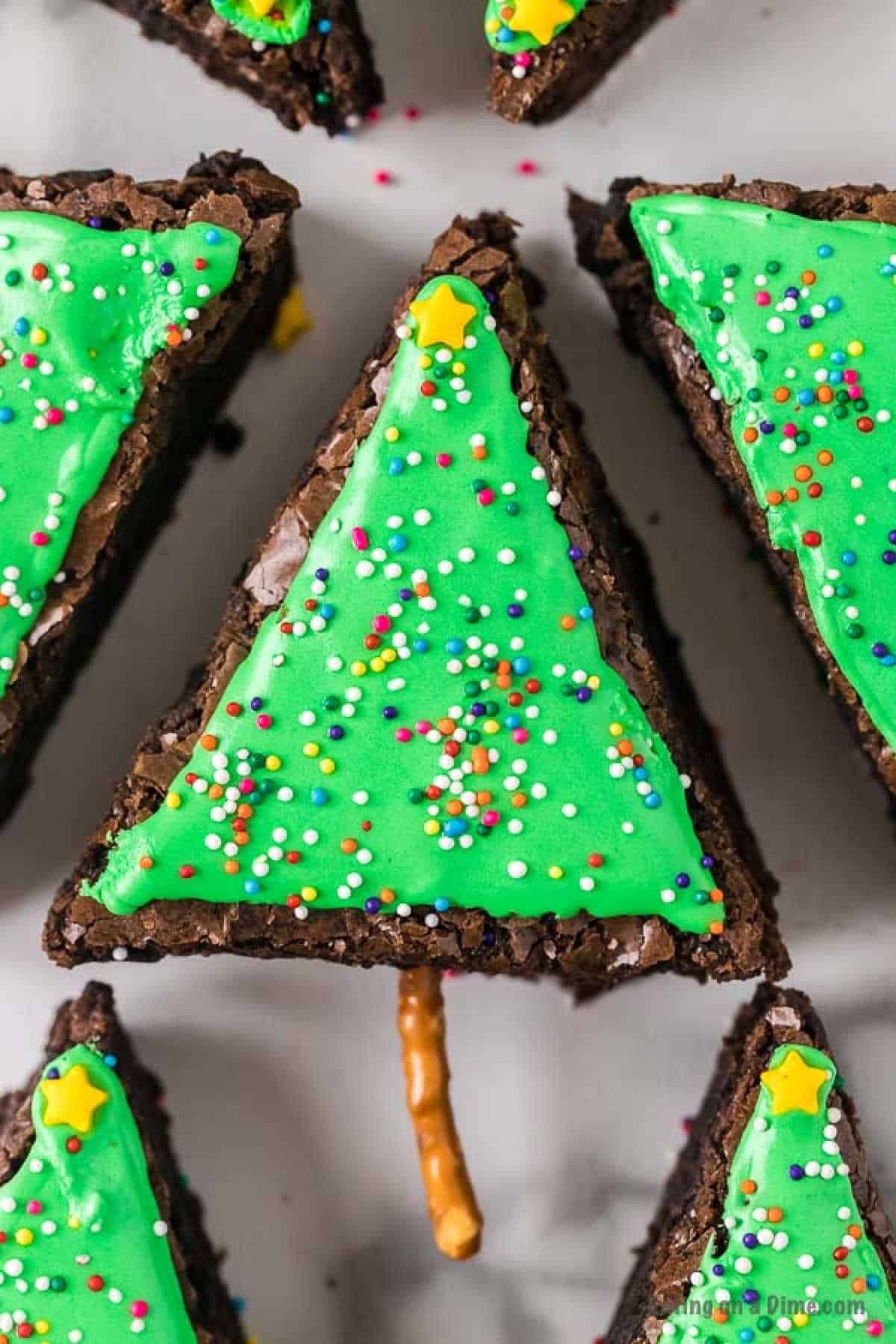 A close-up of Christmas Tree Brownies cut into triangular shapes and decorated to look like festive trees. The brownies are topped with green frosting, colorful sprinkles, and a star-shaped candy. Each brownie has a small pretzel stick as the tree trunk.