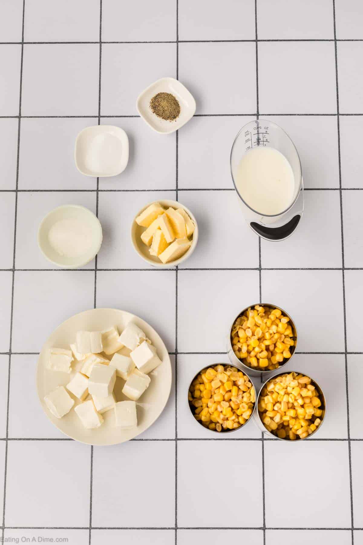 Top-down view of several ingredients on a white tiled surface: butter, flour, canned corn, milk, cream cheese, salt, and pepper. These ingredients appear pre-measured and arranged neatly for preparing a delicious Cream Corn Recipe.