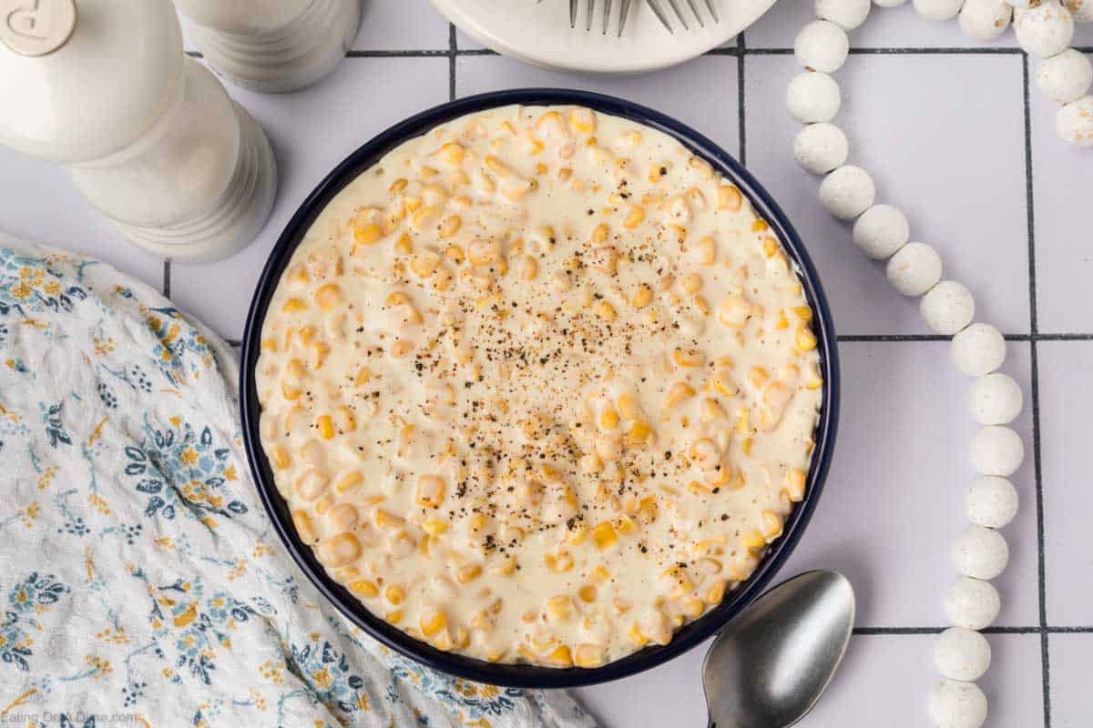 A bowl of creamy corn, highlighting our delicious Cream Corn Recipe, is topped with black pepper and sits on a white tiled surface. Surrounding it are a spoon, a floral cloth, salt and pepper shakers, a white plate with forks, and a string of decorative white beads.