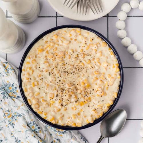 A bowl of creamy corn soup, straight from a cream corn recipe, garnished with black pepper, placed on a tiled surface. Nearby are a floral cloth napkin, a spoon, a set of forks on a plate, salt and pepper shakers, and a string of decorative beads.