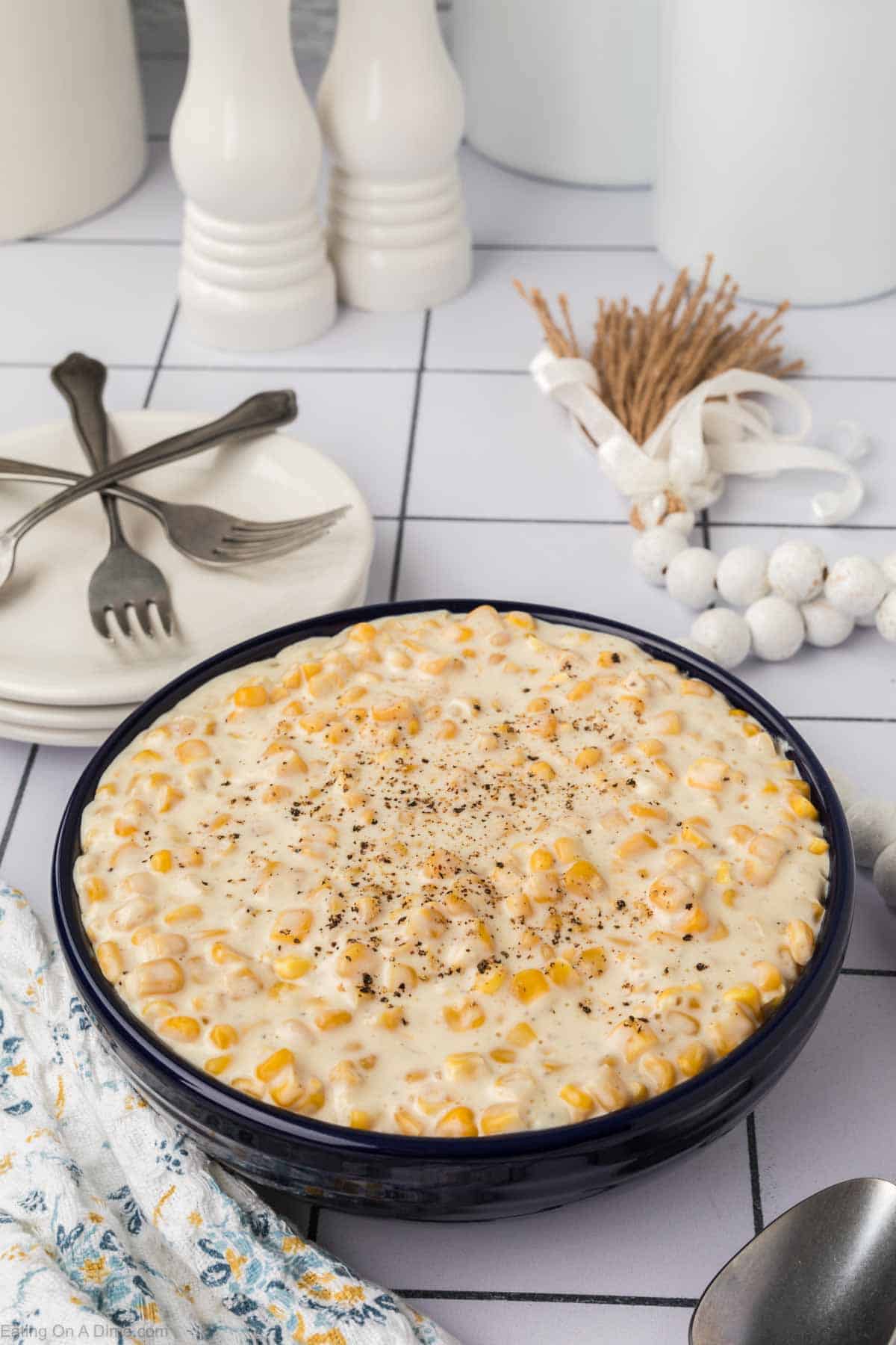 A blue bowl filled with creamy corn, garnished with black pepper, sits on a white tiled surface. This Cream Corn Recipe is accompanied by three forks on stacked white plates, a patterned napkin, a bundle of wheat, and a pair of white ceramic shakers.