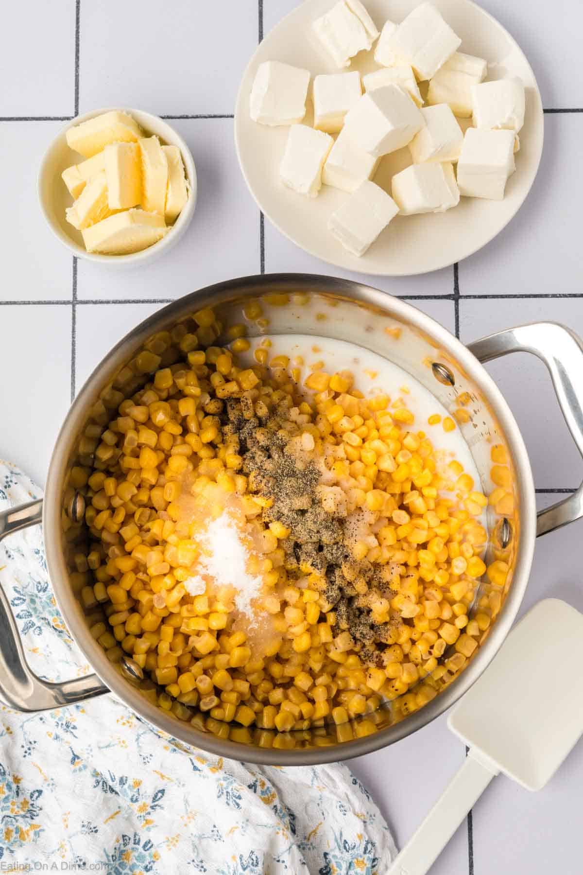 A top-view photo showcases a Cream Corn Recipe: a cooking pot filled with corn, cream, and seasonings. Nearby, there's a small bowl of butter, a plate with cream cheese cubes, and a floral-patterned cloth. A white spatula rests on the counter next to the pot.