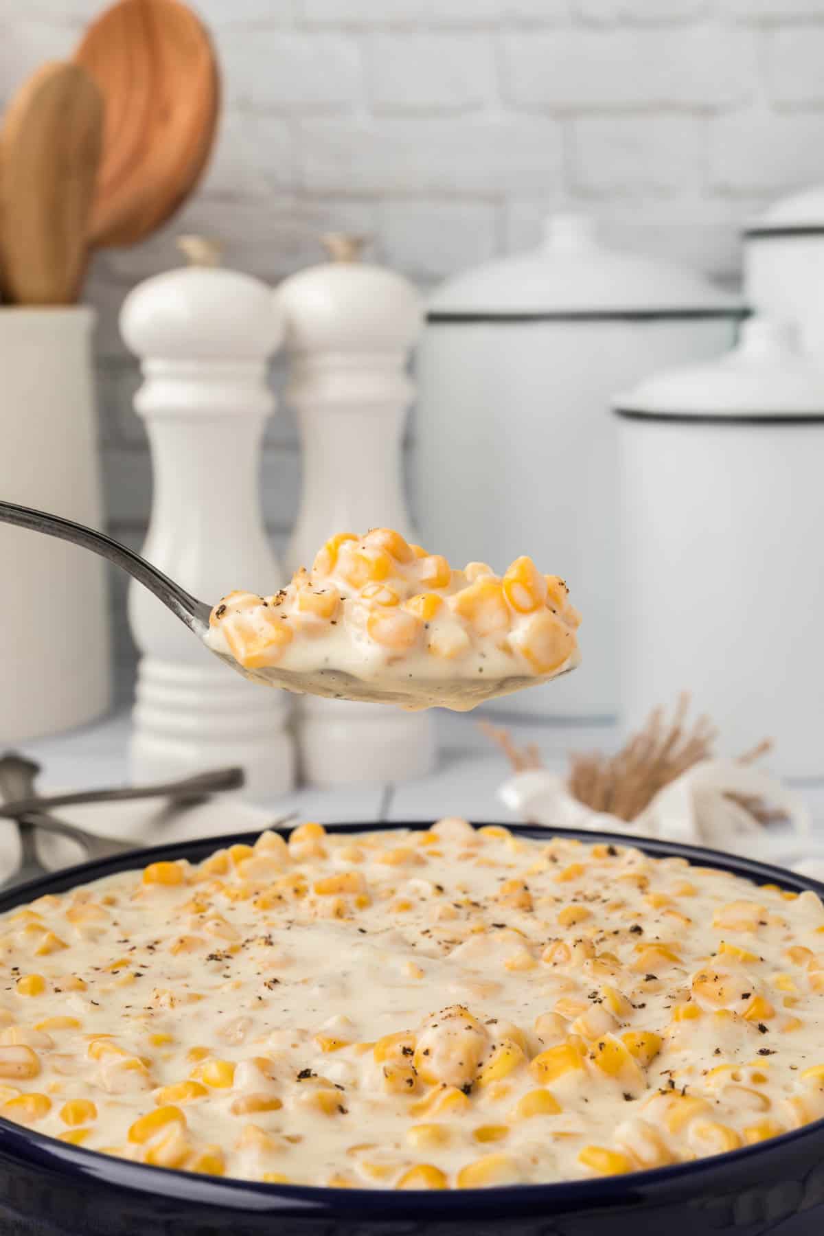 A close-up of a spoonful of creamy corn being held above a dish filled with the same delightful cream corn recipe. The dish rests on a kitchen counter, surrounded by white ceramic containers, wooden utensils, and white salt and pepper shakers in the subtly blurred background.