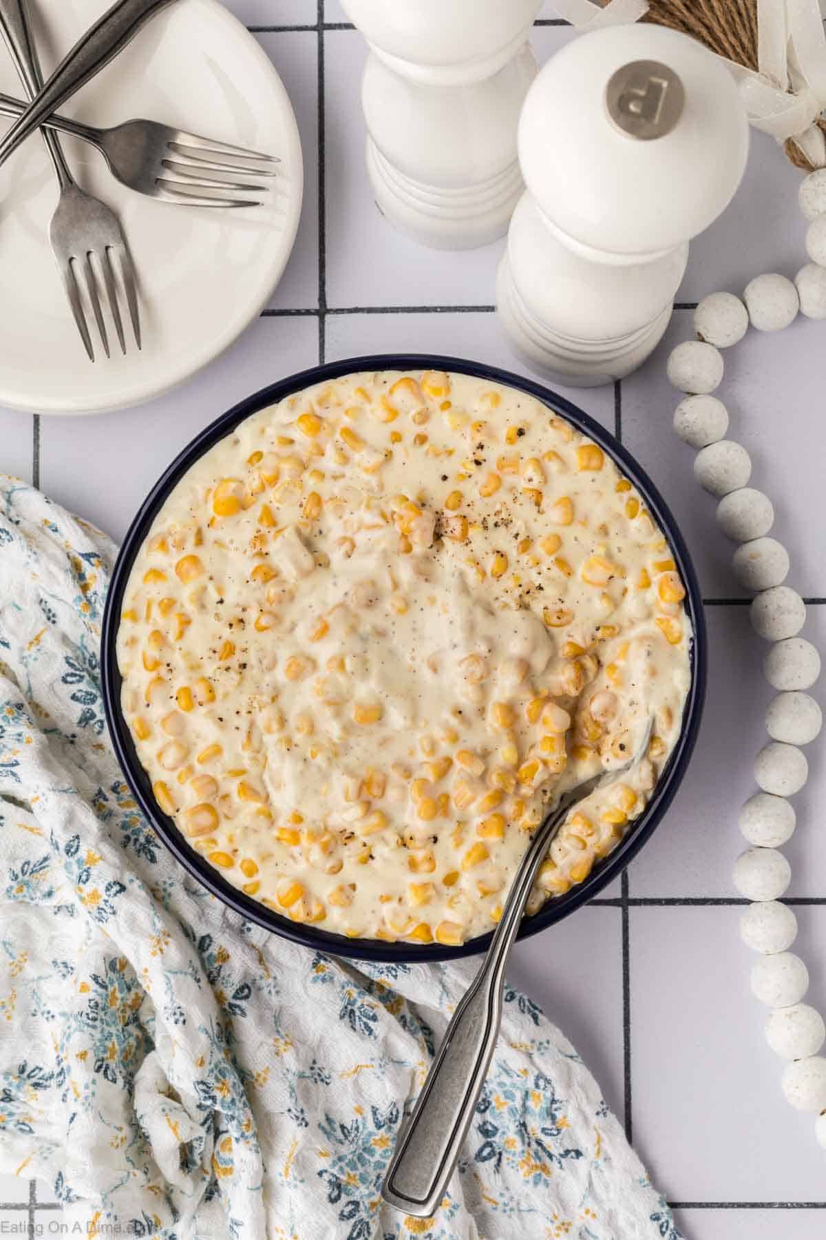 A bowl of creamy corn casserole, made from a delightful cream corn recipe, is topped with pepper and set on a white tiled surface. A spoon rests in the bowl. Surrounding items include a floral cloth, white pepper and salt shakers, a beaded decoration, and a white plate with a fork and spoon.