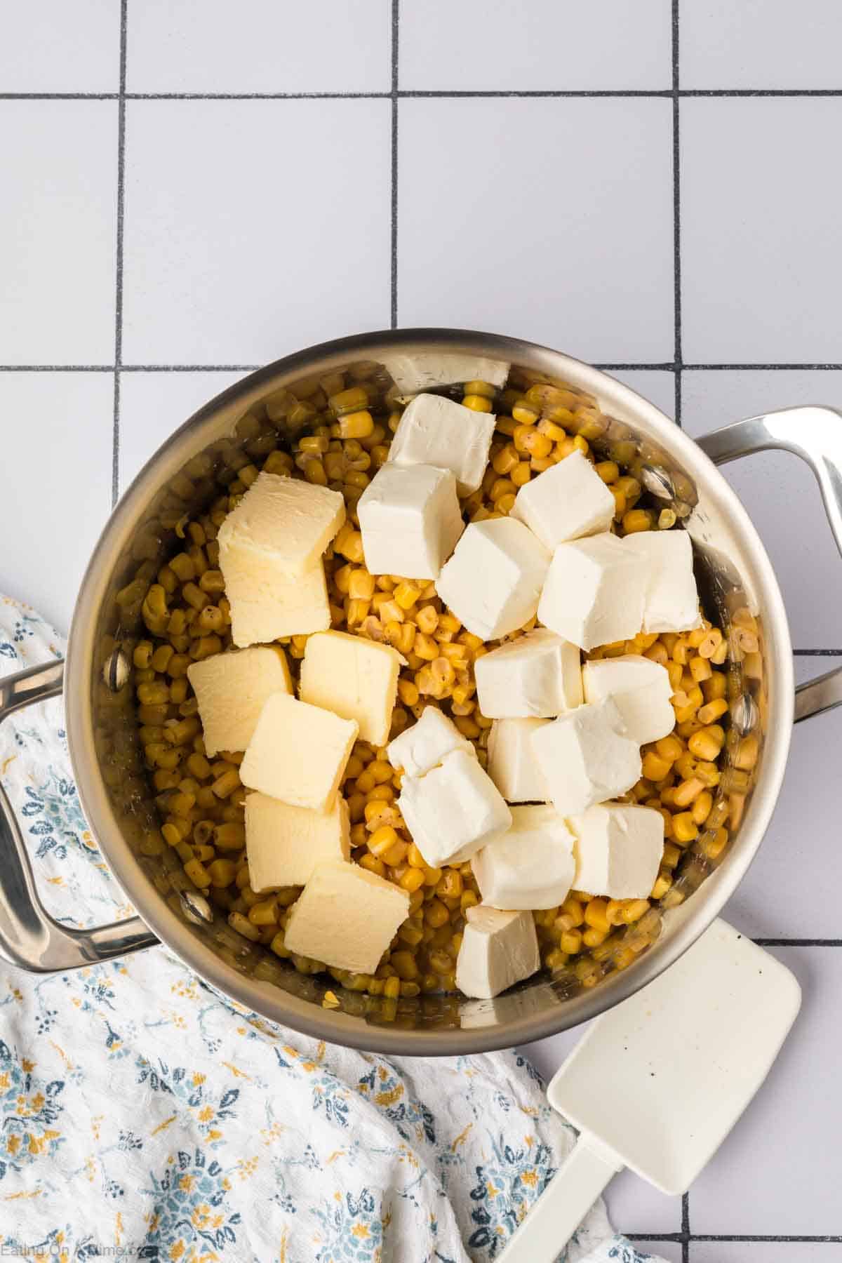 A pot filled with corn kernels, topped with several cubes of cream cheese and butter, sits on a tiled surface. Next to it, a patterned cloth is partially visible with a white spatula resting on top, ready to stir up this delectable cream corn recipe.