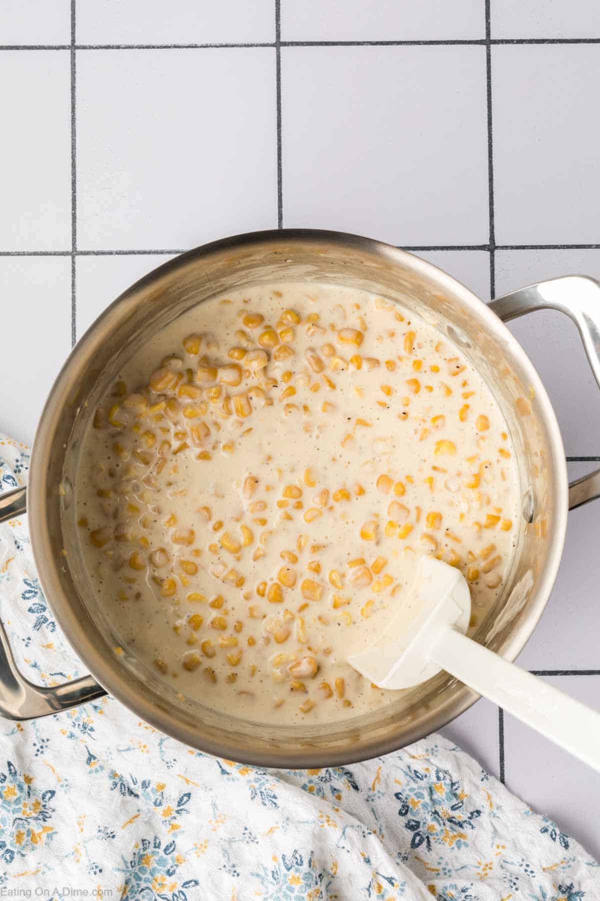 A metal pot containing creamy, cooked corn with a white sauce sits invitingly. A white spatula is nestled inside, ready for the next stir. The pot is placed on a grid-patterned surface, with a glimpse of floral-patterned cloth on the left side—a perfect setting for your favorite cream corn recipe.