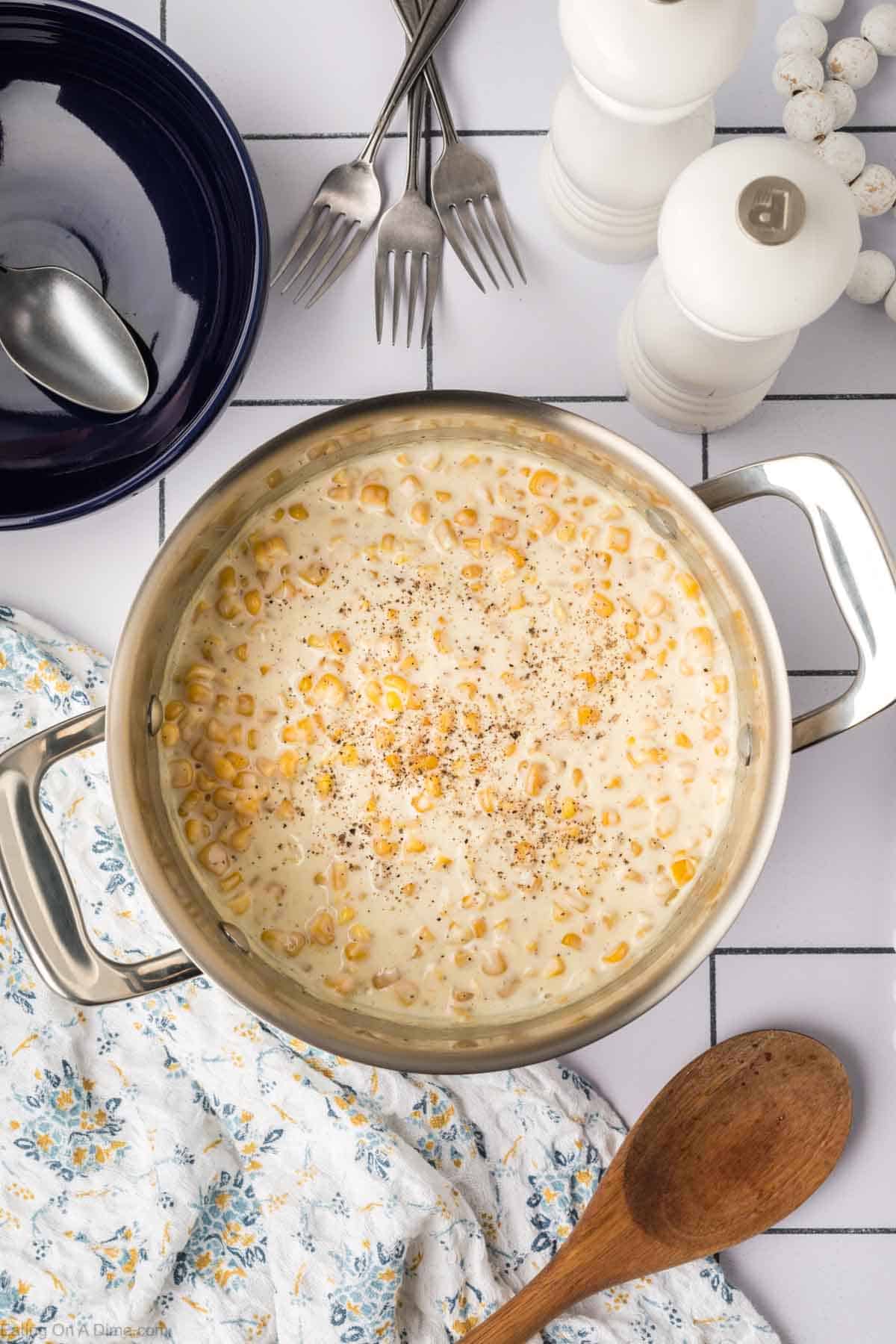 A pot of creamy corn chowder, inspired by a cherished cream corn recipe, topped with freshly ground black pepper sits on a tiled countertop. Nearby are a wooden spoon, a floral-patterned cloth, a black bowl, silver forks and spoons, and two white salt and pepper shakers.