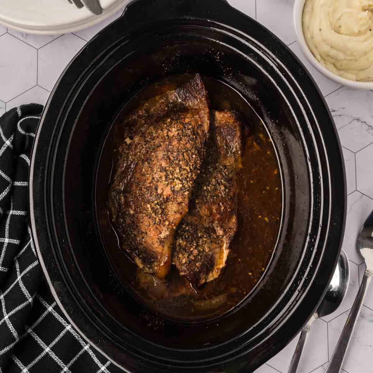 Close-up of a slow cooker containing cooked, seasoned pork resting in a rich, dark broth, reminiscent of a crockpot steak recipe. It's placed on a hexagonal patterned countertop next to a black and white checkered cloth, silver cutlery, and a bowl of creamy mashed potatoes.