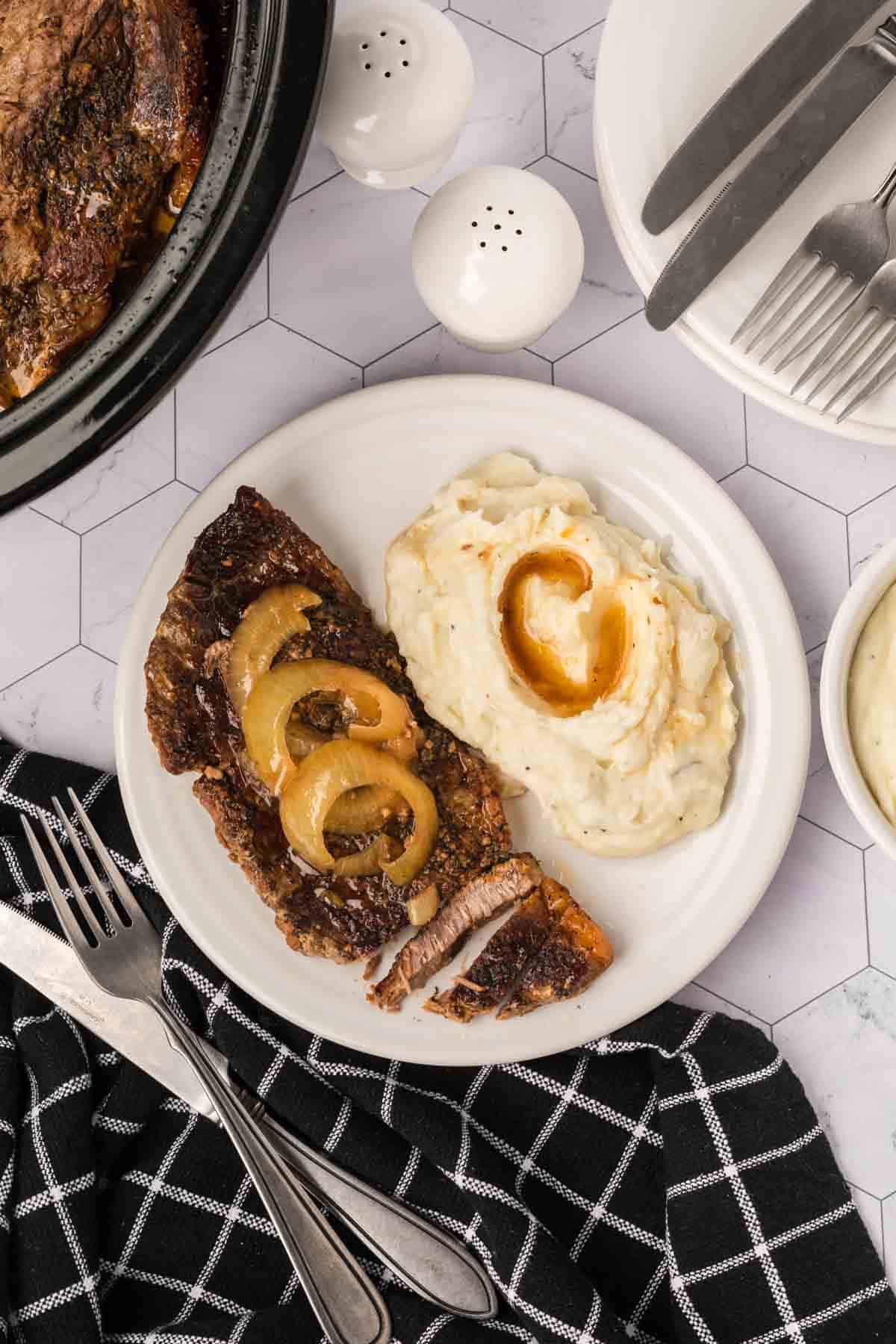 A plate with crockpot steak slices topped with onion rings and a serving of mashed potatoes and gravy. Beside it are a black and white checkered napkin, fork, knife, and salt and pepper shakers on a geometric-patterned surface.