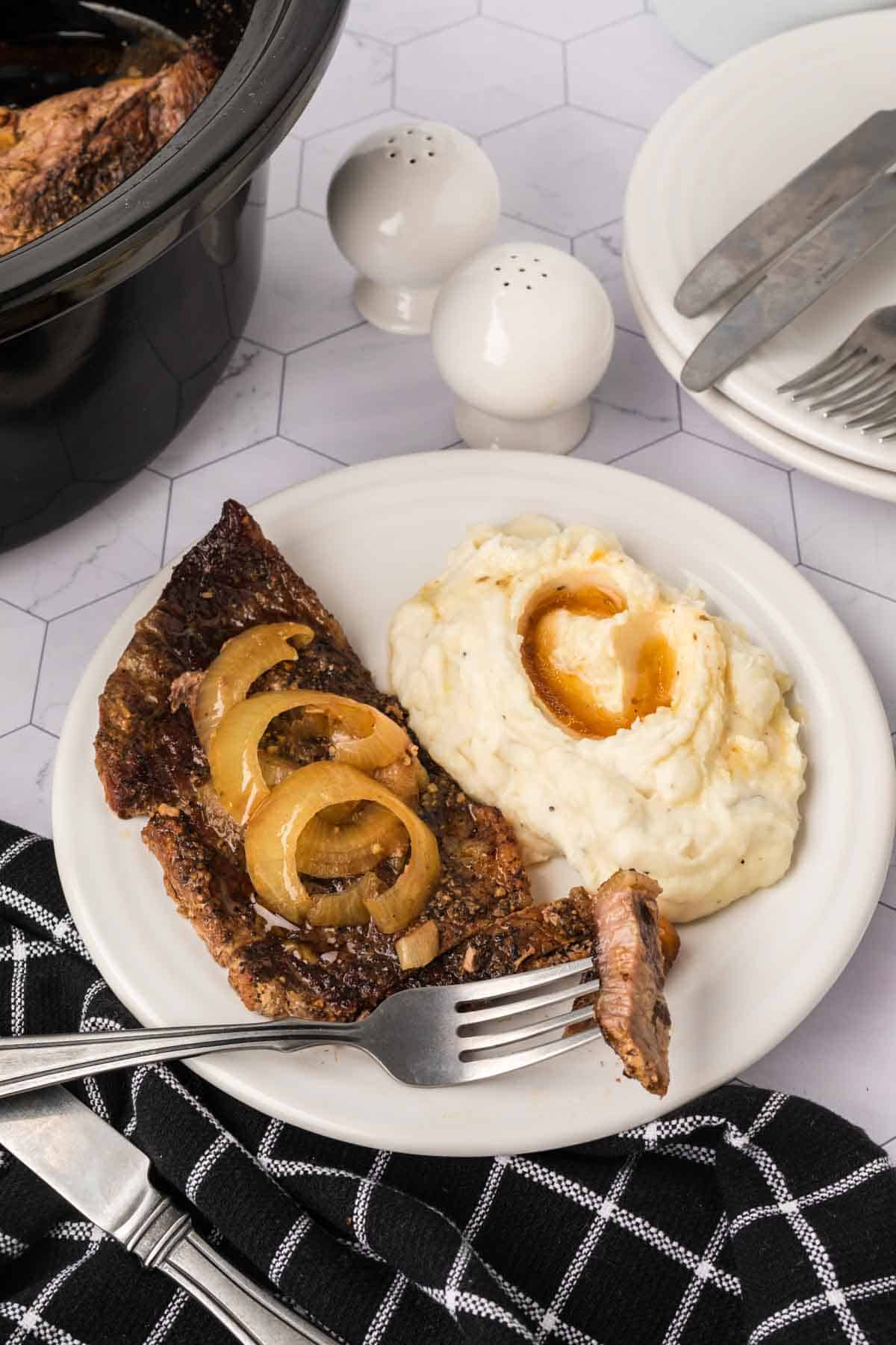 A plate featuring a slice of roast beef topped with cooked onions, reminiscent of a savory crockpot steak recipe, is accompanied by creamy mashed potatoes with a pat of butter. A fork rests on the plate, while a black and white checkered cloth, cutlery, and salt and pepper set lie nearby.