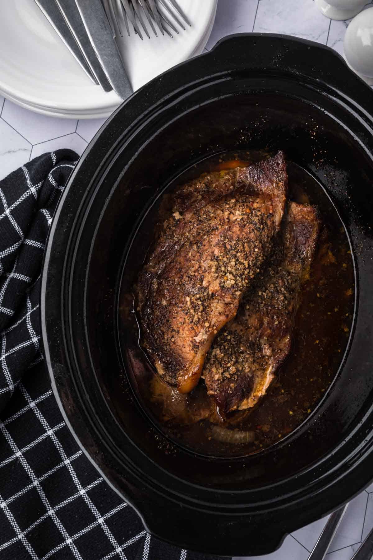 Slow-cooked beef with a brown, seasoned crust sits invitingly in a black crockpot, showcasing the perfect crockpot steak recipe. It's placed on a countertop with a black and white checkered kitchen towel beside it, accompanied by a stack of white plates and shiny silver forks.