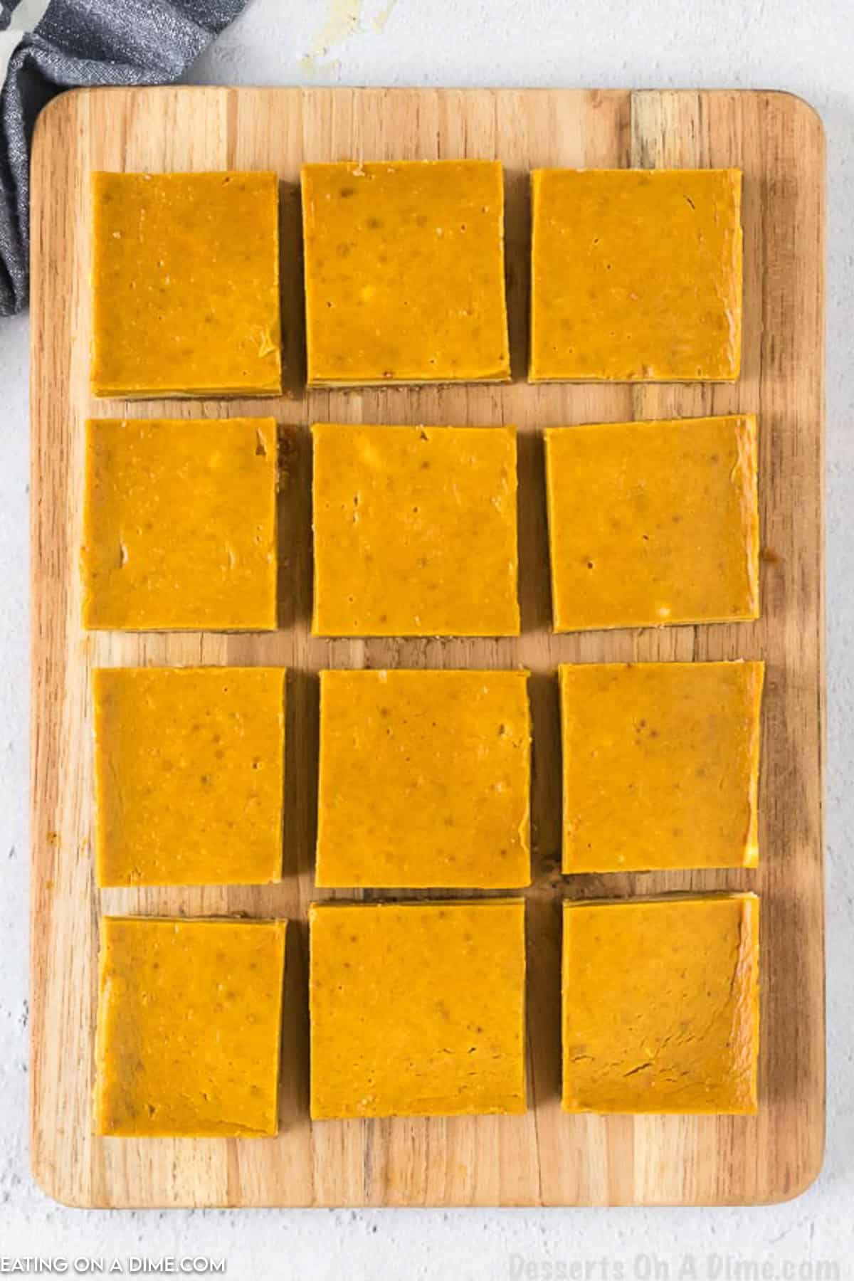 A wooden cutting board with twelve neatly arranged square pieces of pumpkin fudge. The fudge squares are evenly cut and have a smooth, rich orange appearance, reminiscent of Pumpkin Cheesecake Bars. A dark cloth napkin partially extends onto the corner of the cutting board.