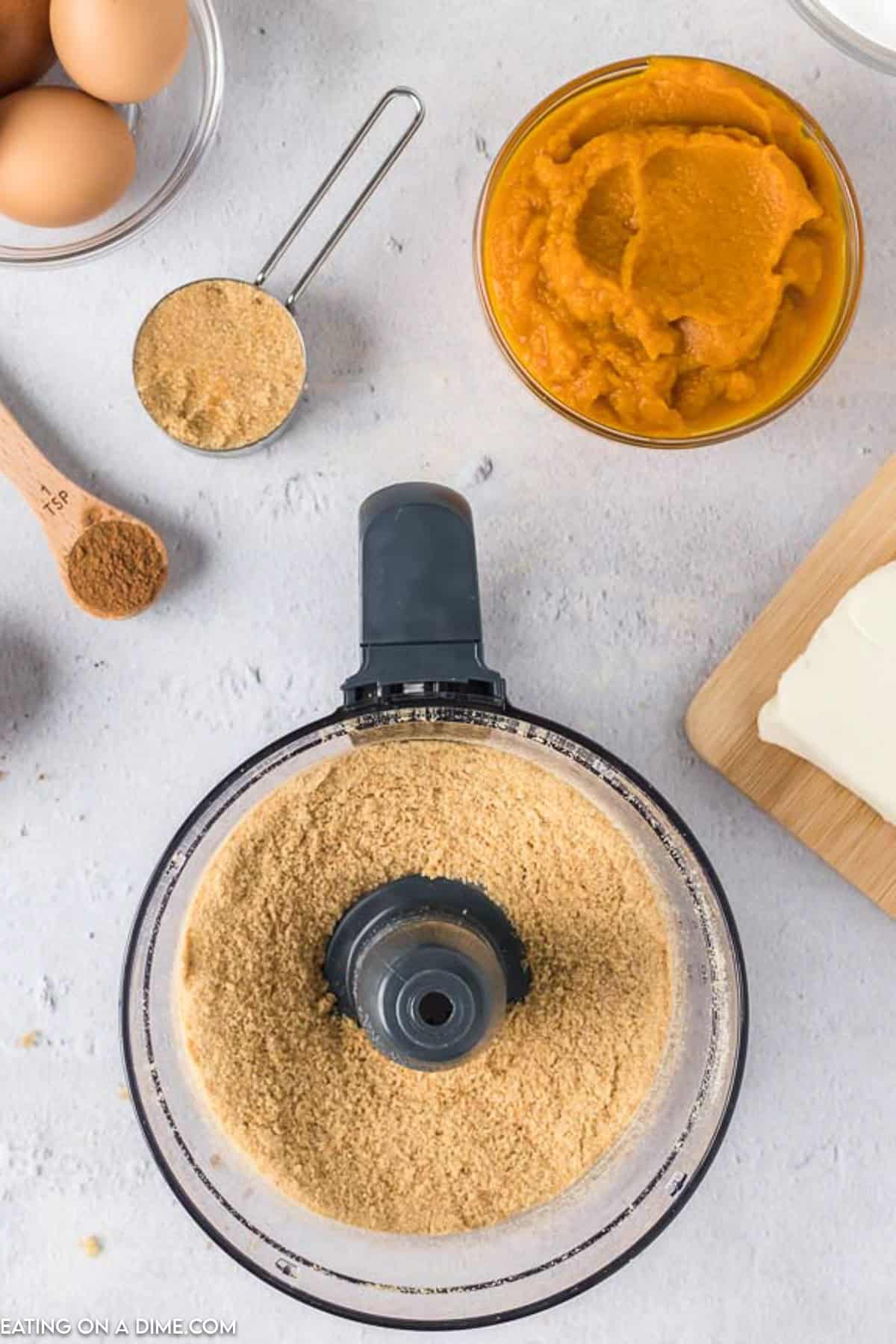 A top-view photo shows baking ingredients spread around a food processor with ground crumbs. Nearby are bowls containing pumpkin puree and brown sugar, a measuring spoon with cinnamon, a block of cream cheese on a cutting board, and three eggs—everything you need to create delicious Pumpkin Cheesecake Bars.