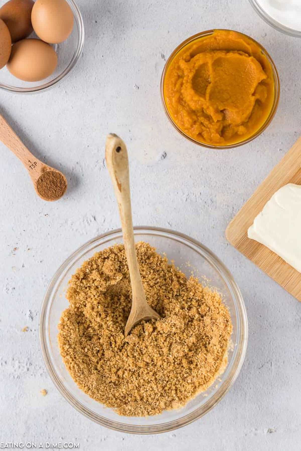 A bowl of crushed graham crackers with a wooden spoon, a small bowl of pumpkin puree, cream cheese on a wooden cutting board, eggs, and a measuring spoon with spices are arranged on a light gray surface, suggesting the preparation for Pumpkin Cheesecake Bars.