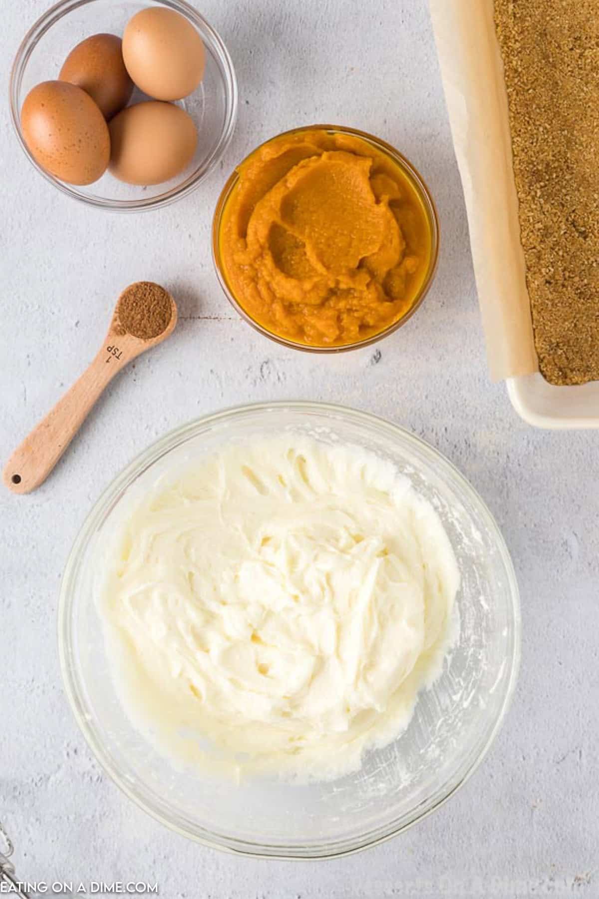 An overhead view of baking ingredients for Pumpkin Cheesecake Bars on a white countertop. There are four brown eggs, a wooden spoon with a small amount of spice, a bowl of pumpkin puree, a bowl of whipped cream cheese mixture, and a baking tray with a crumb crust.