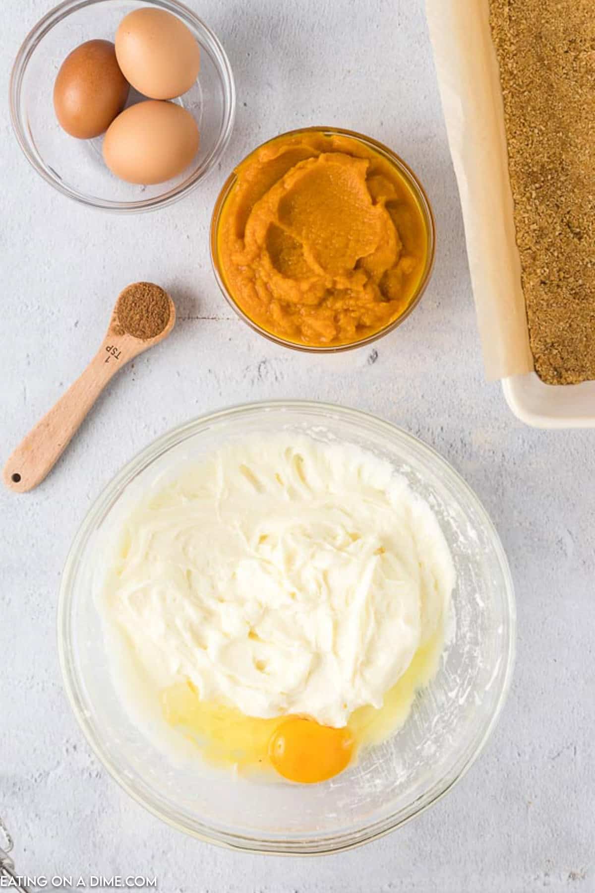 A bowl of whipped cream cheese with an egg on top, beside a bowl of pumpkin puree, a wooden spoon with cinnamon, and three whole eggs. A rectangular baking dish with a graham cracker crust for Pumpkin Cheesecake Bars is shown on the side. The items are on a light-colored surface.